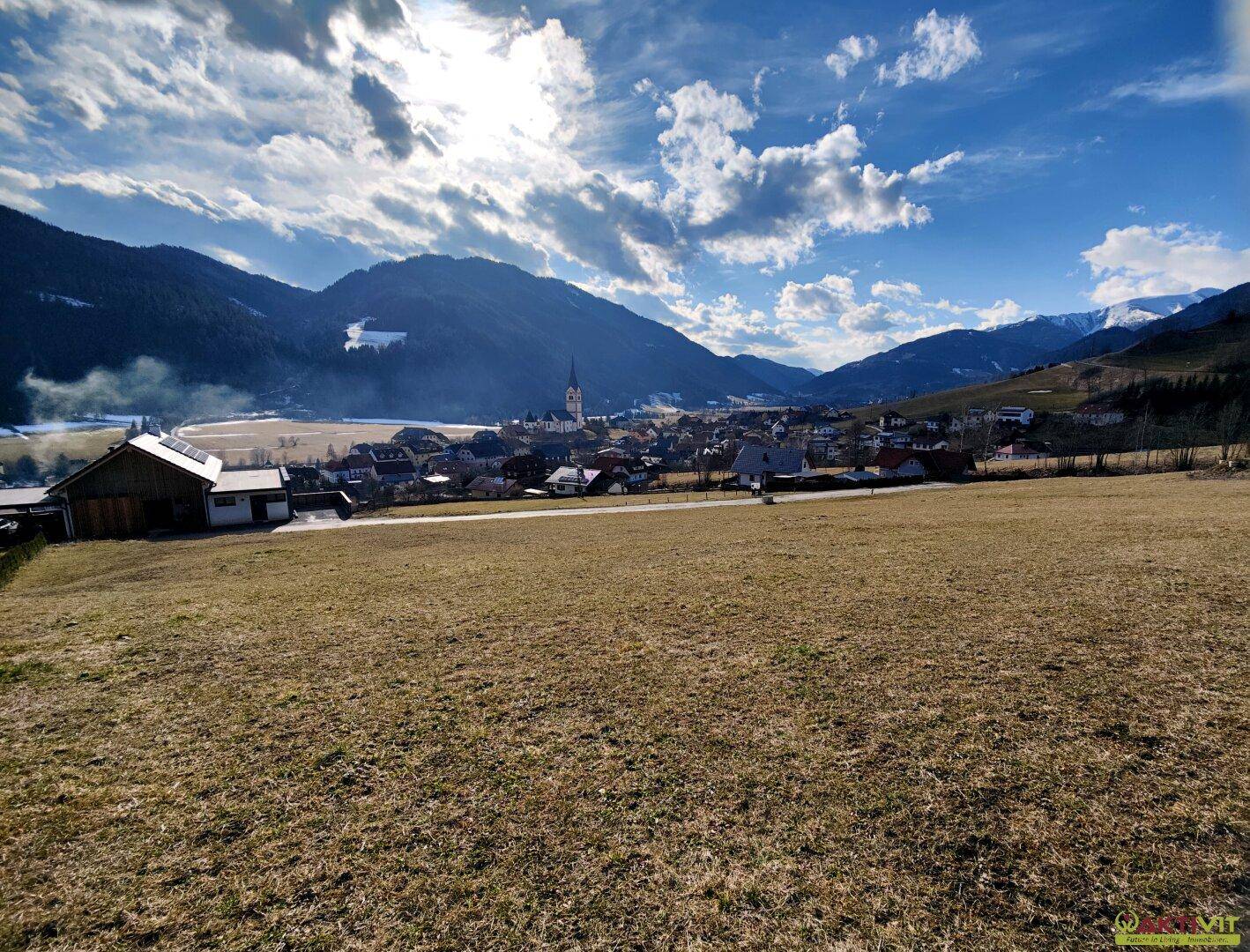 Panoramablick über ein Tal mit einem Dorf, Kirche und schneebedeckten Bergen im Hintergrund.