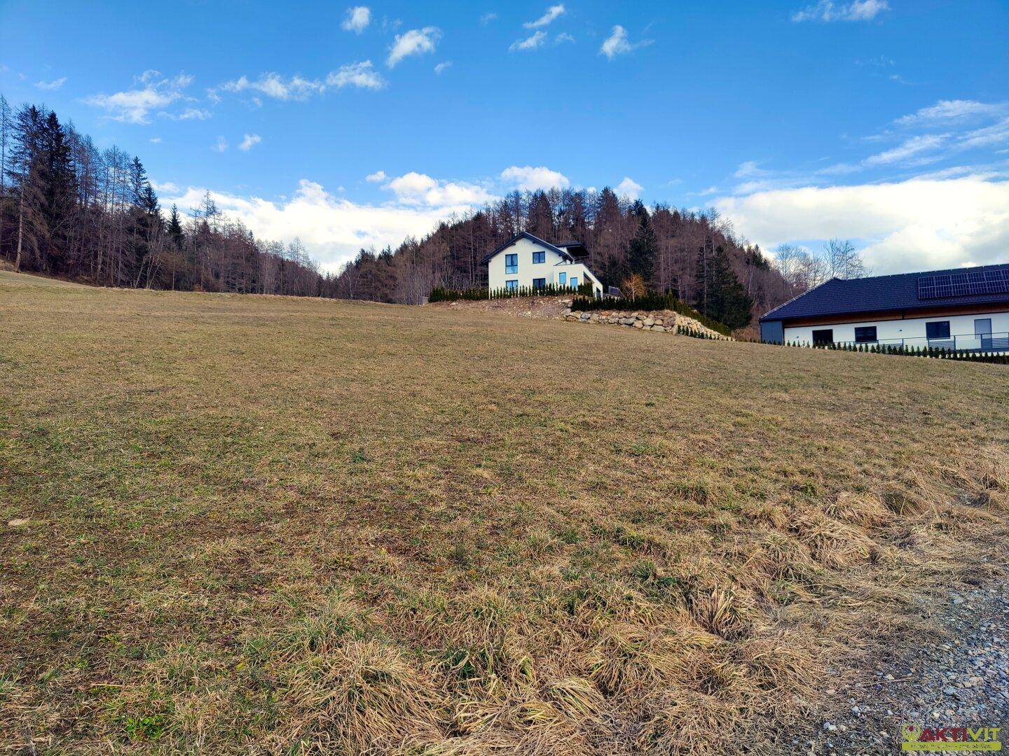 Offenes Feld mit Blick auf ein modernes Haus und bewaldete Hänge unter blauem Himmel.