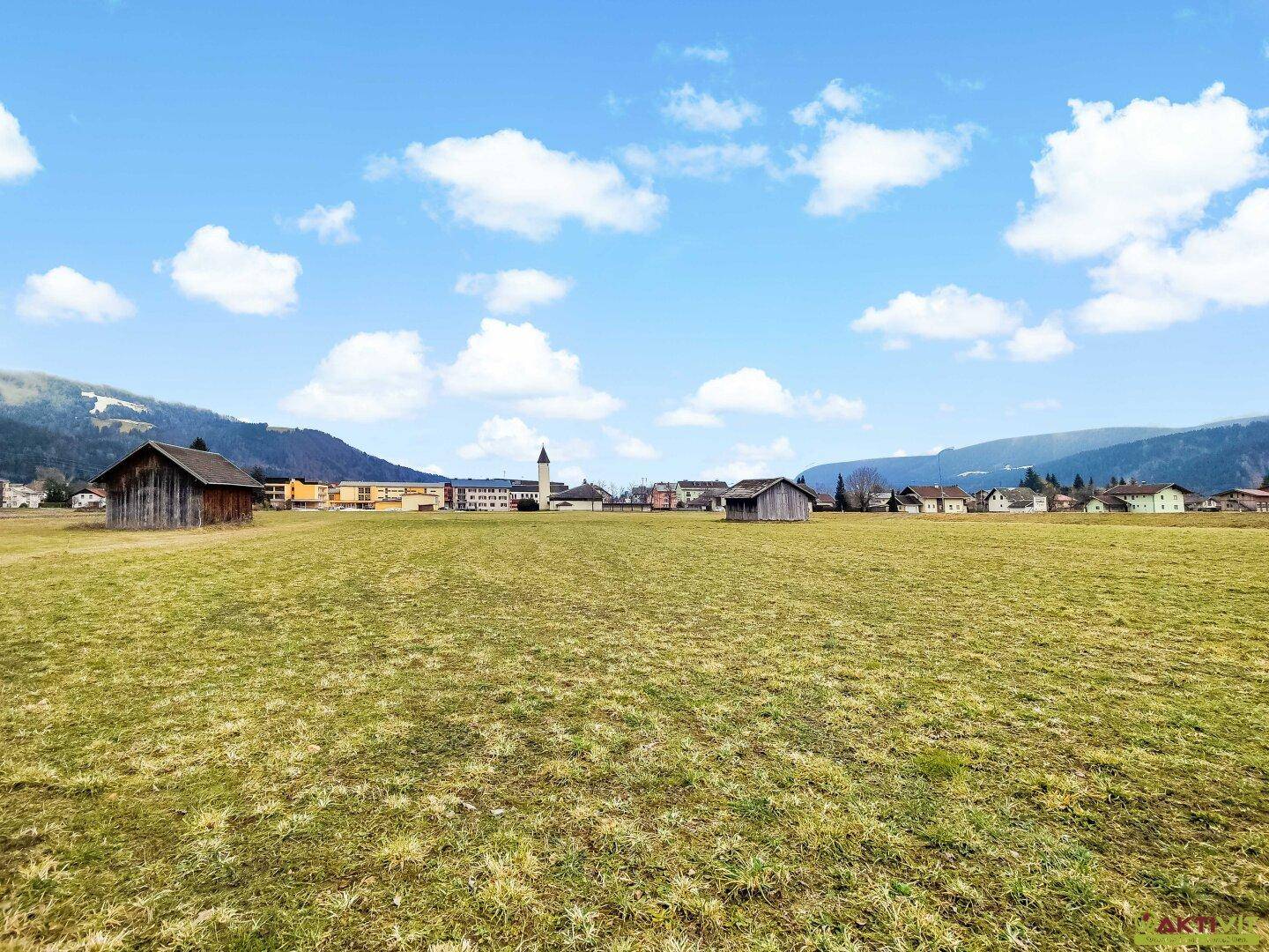 Offenes Feld mit Blick auf eine kleine Ortschaft und die sanften Hügel der Umgebung unter blauem Himmel.