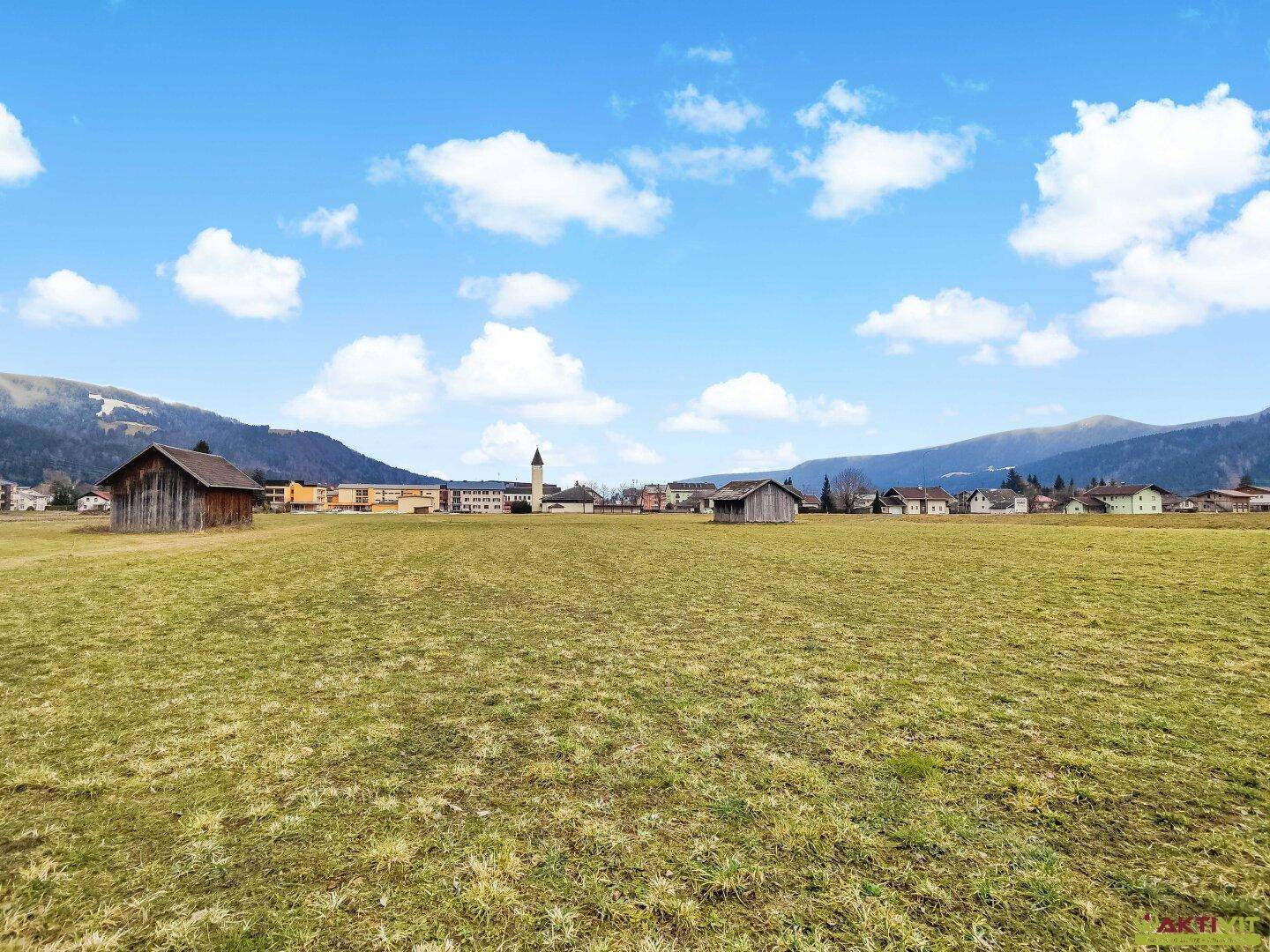 Weitläufige Wiesenfläche mit Fernblick auf ein Dorf und die bewaldeten Berge im Hintergrund bei sonnigem Wetter.