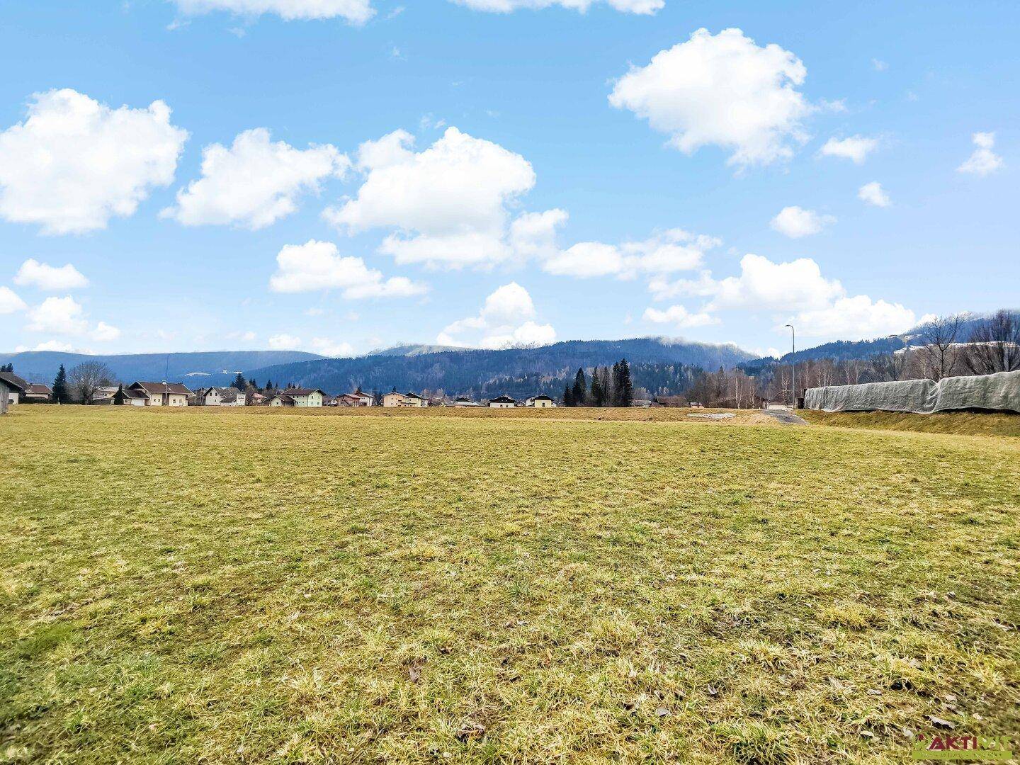 Großes Feld mit Blick auf eine Häuserreihe und bewaldete Berge am Horizont unter einem klaren Himmel.