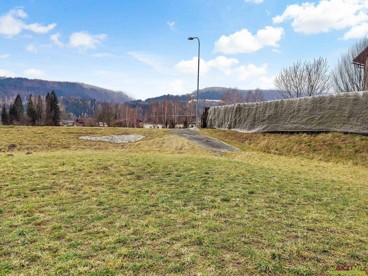 Grünfläche mit einem Laternenpfahl, einer Abdeckung am Rand und bewaldeten Bergen im Hintergrund.