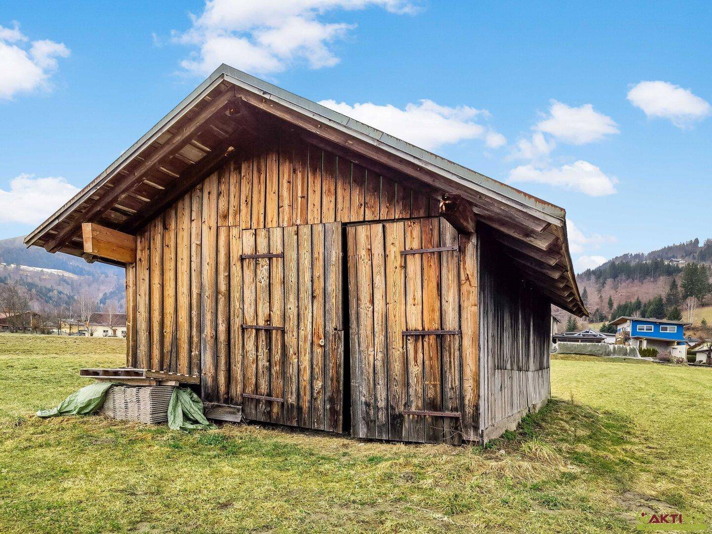 Rustikaler Holzschuppen mit Doppeltüren auf einer grünen Wiese unter einem strahlend blauen Himmel.