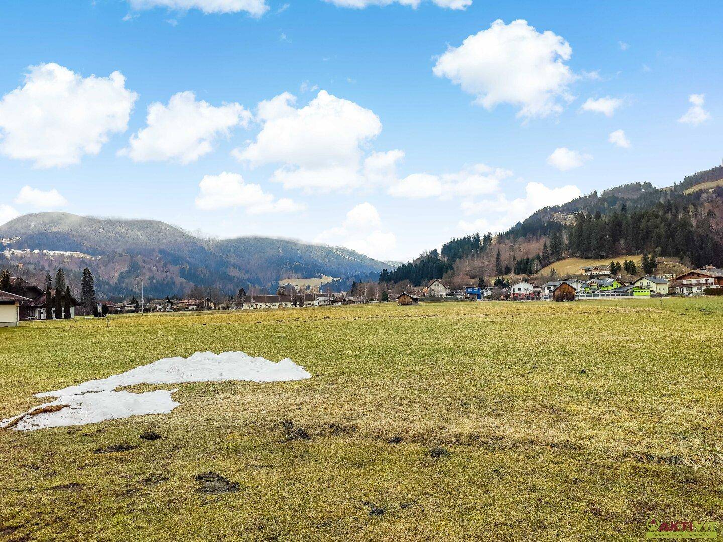 Weite Wiesenfläche mit einem Schneefleck, umgeben von Häusern und bewaldeten Bergen in der Ferne.