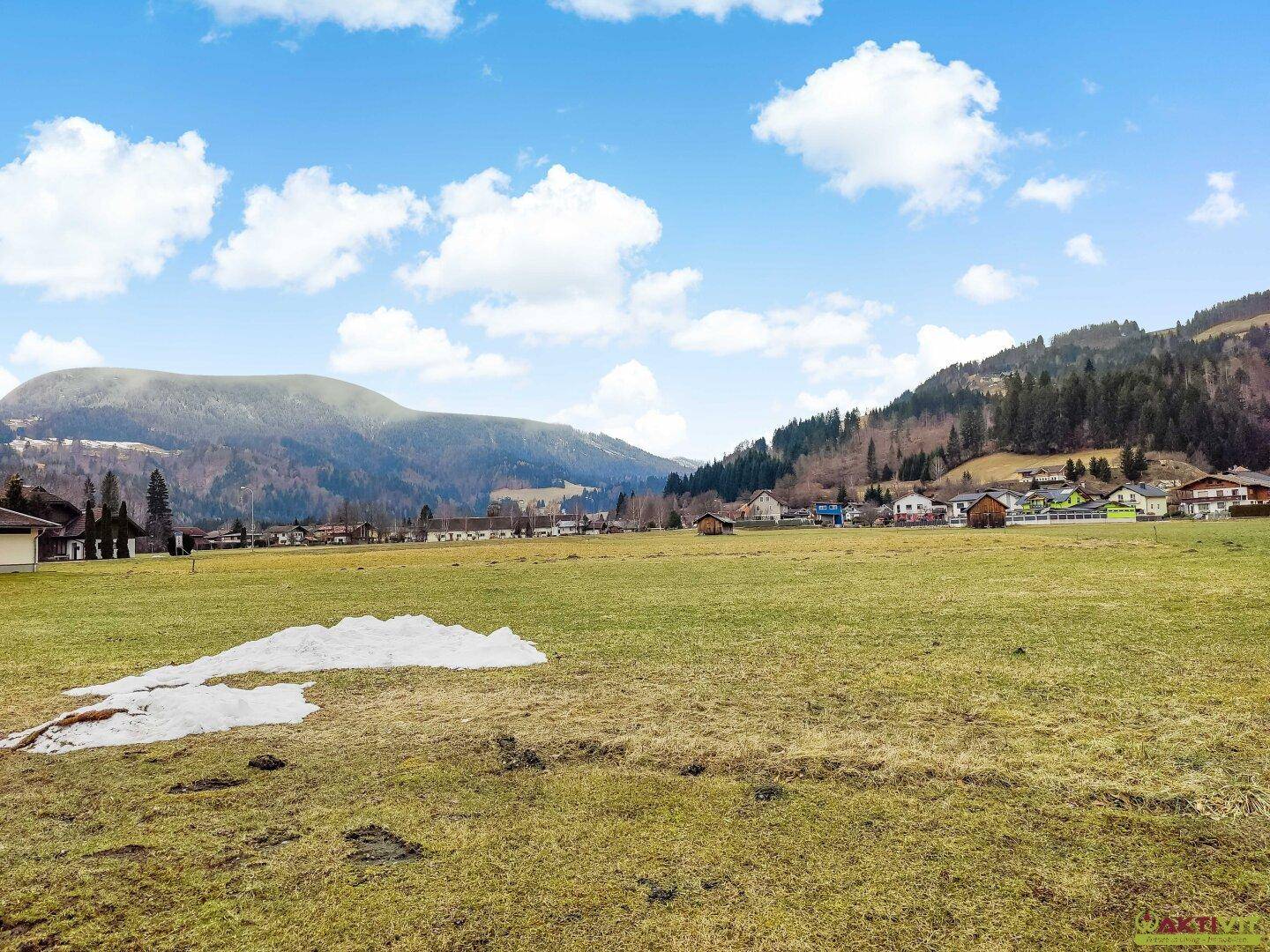 Offenes Feld mit Restschnee, umgeben von einer ländlichen Bebauung und bewaldeten Hügeln am Horizont.