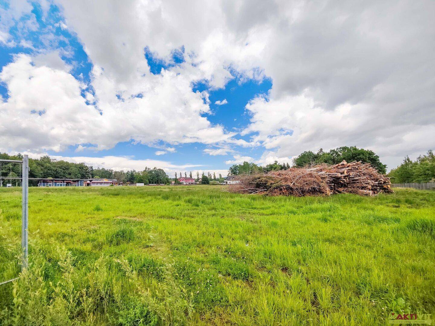 Weitläufiges grünes Feld mit einem großen Holzstapel und Gebäuden sowie Bäumen in der Ferne.