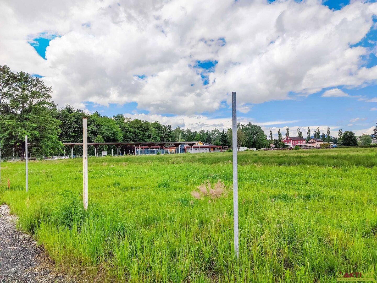 Weitläufiges grünes Feld mit zwei Metallpfosten im Vordergrund und Gebäuden sowie Bäumen in der Ferne.