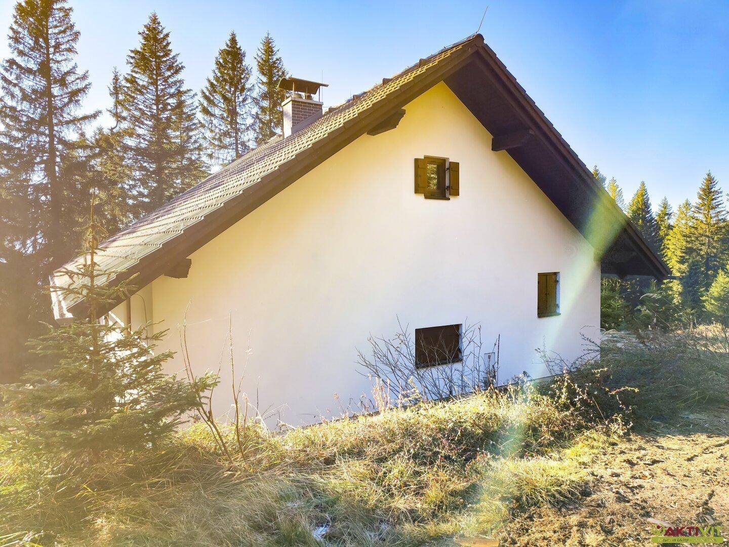 Seitenansicht des Hauses mit weißer Fassade und kleinen Fenstern, eingebettet in die Natur.