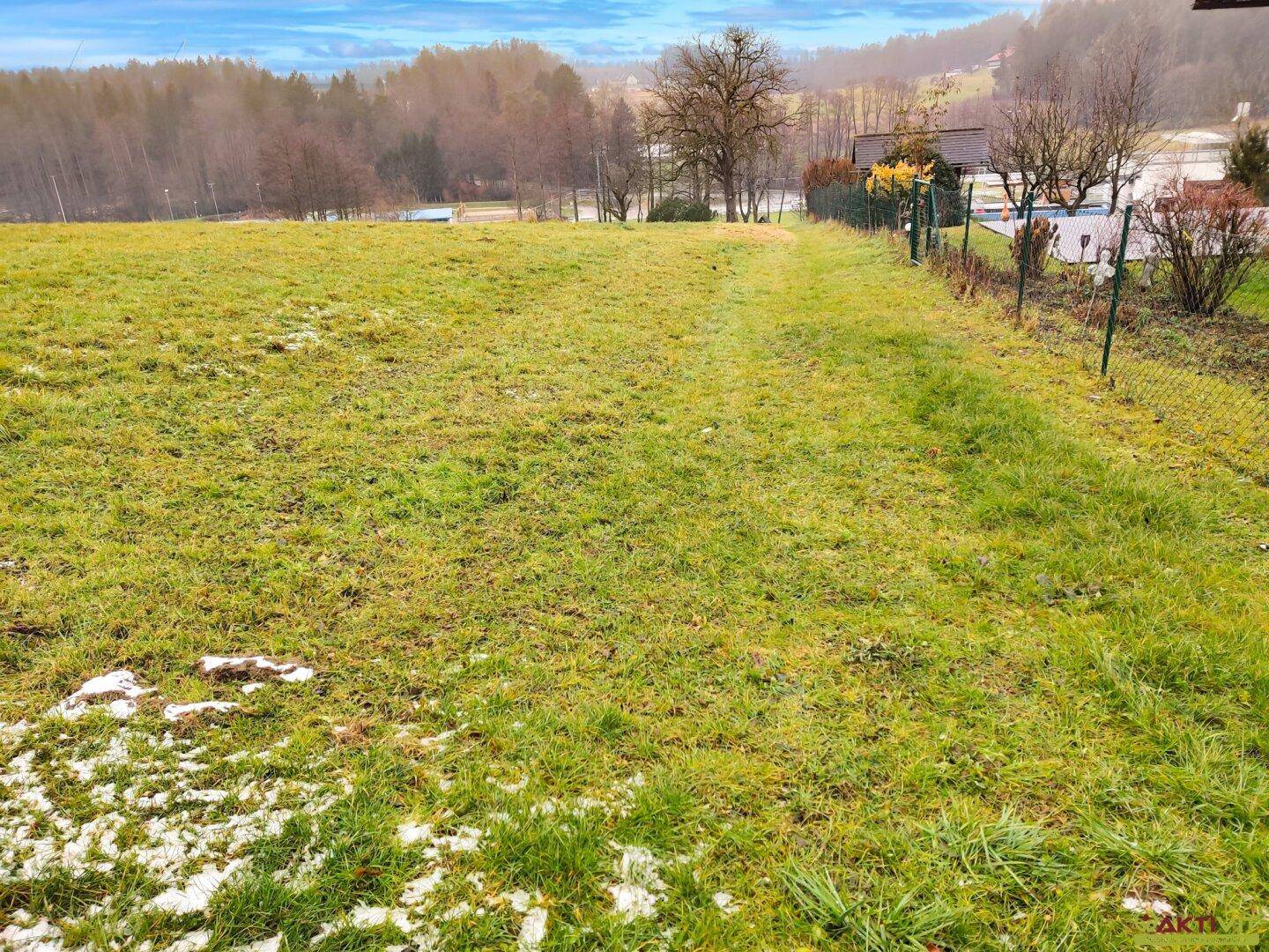 Grünes Grundstück mit Zaun und vereinzelten Schneeflecken, umgeben von ländlicher Natur.