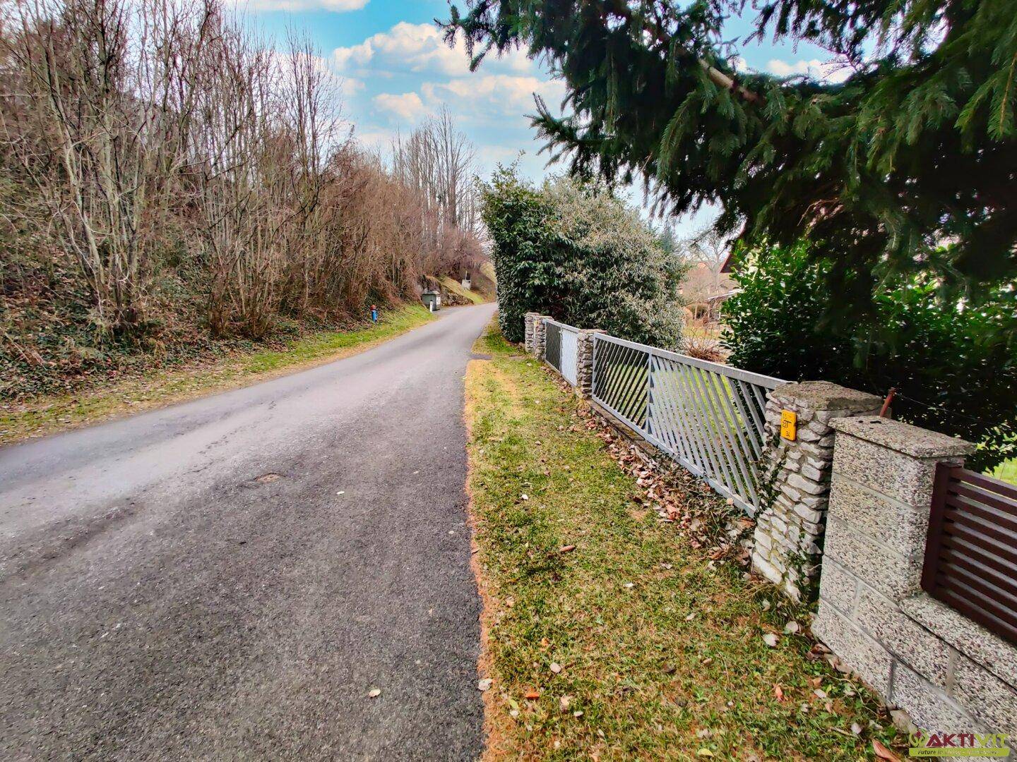 Ländliche Straße mit angrenzendem Grundstück und Blick auf die bewaldete Hügellandschaft.
