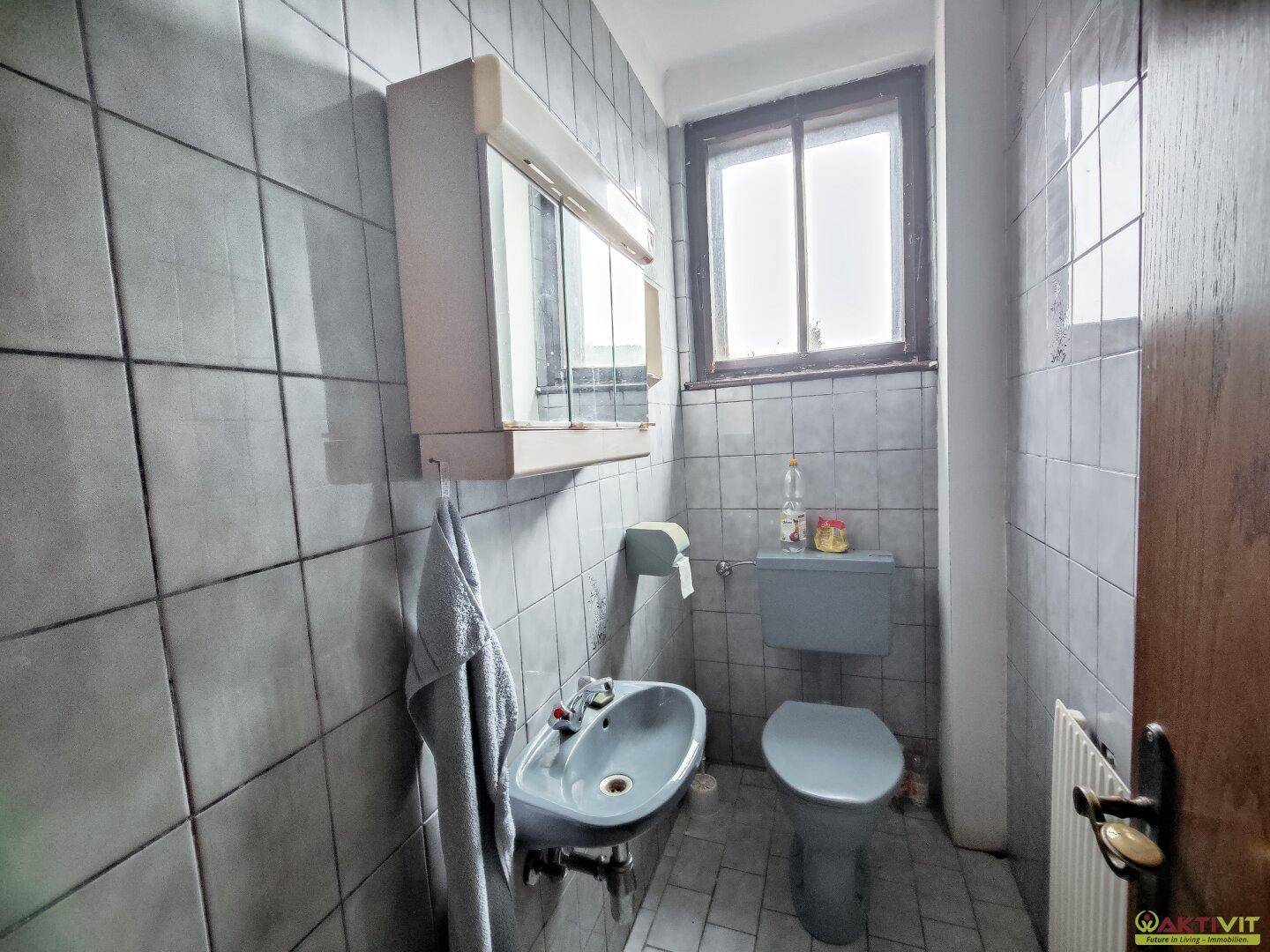 Kleines WC mit Fenster, Waschbecken und grauen Fliesen.