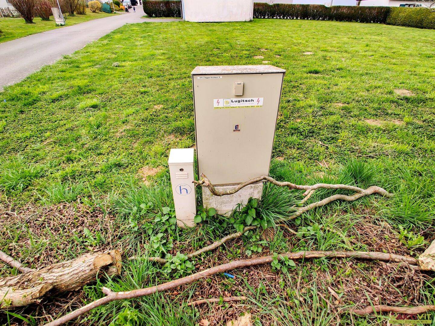 Ein Versorgungsverteilerkasten steht auf einem grasbewachsenen Streifen neben einem gepflasterten Weg.