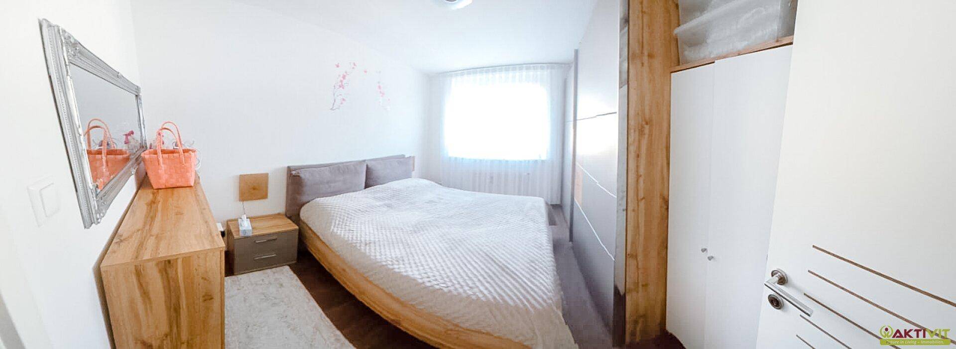 Schlafzimmer mit hellem Holzbett, Spiegel und weißem Kleiderschrank, gut beleuchtet durch ein Fenster.