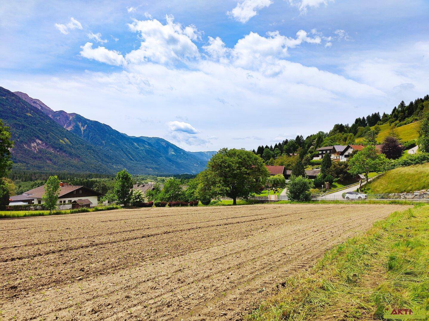 Gepflügtes Feld mit Häusern am Hang und einer weiten Berglandschaft im Hintergrund.
