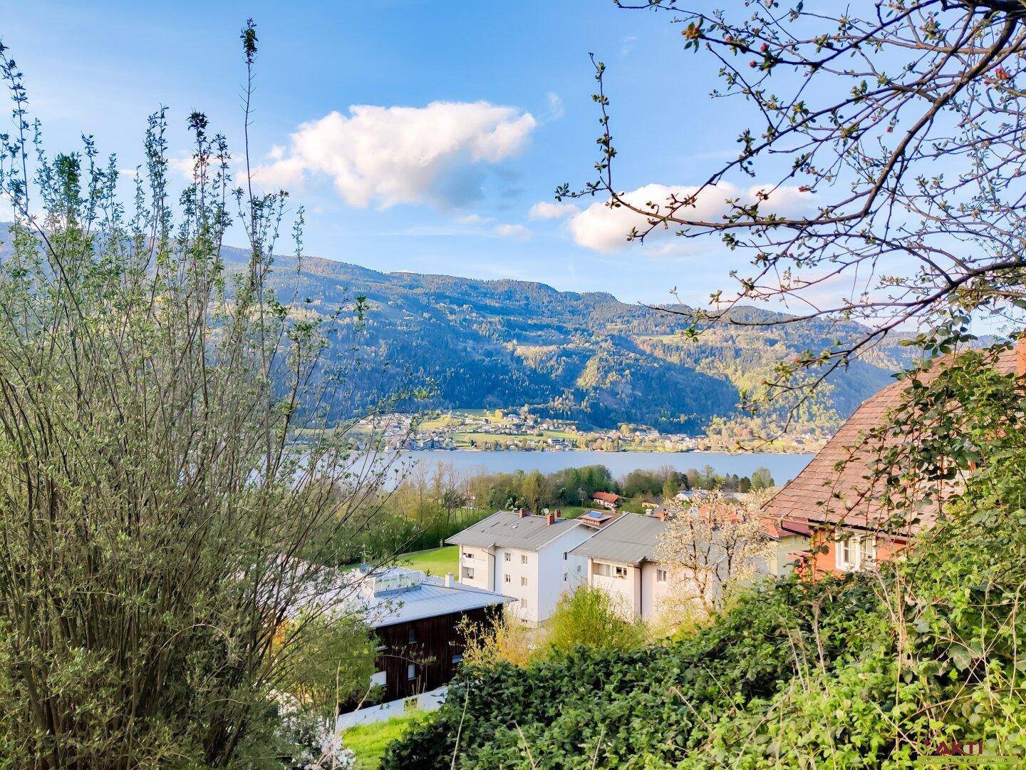Weitläufiger Blick über den See und die Berge mit Siedlungshäusern im Vordergrund unter blauem Himmel.