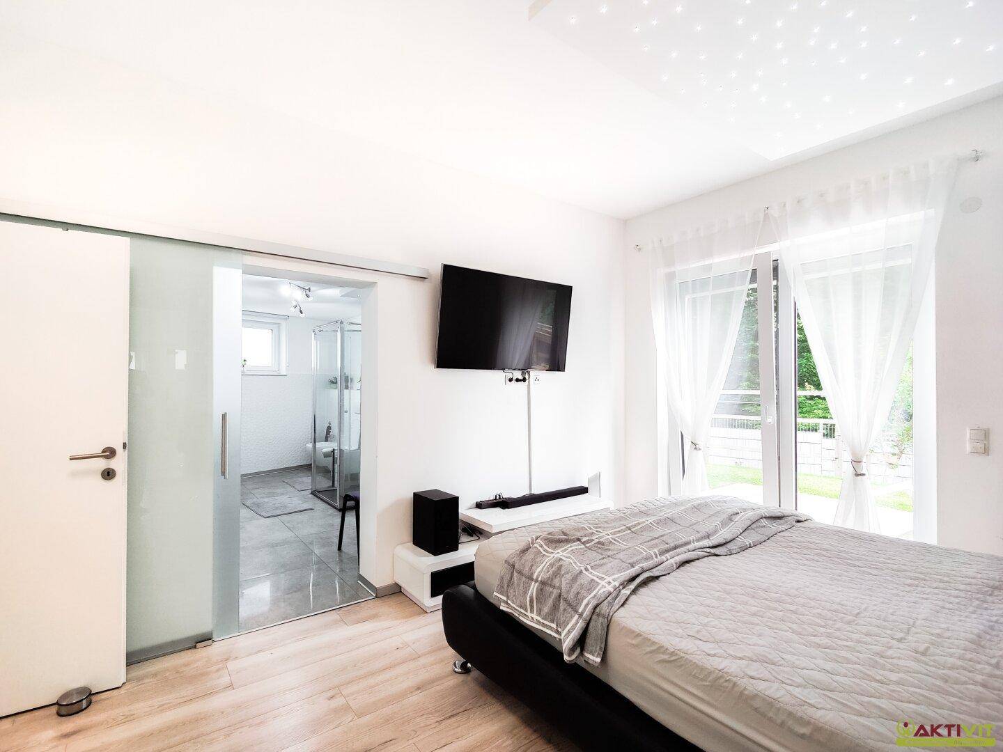 Schlafzimmer mit Holzboden, Terrassenzugang und Schiebetür zum modernen Bad.