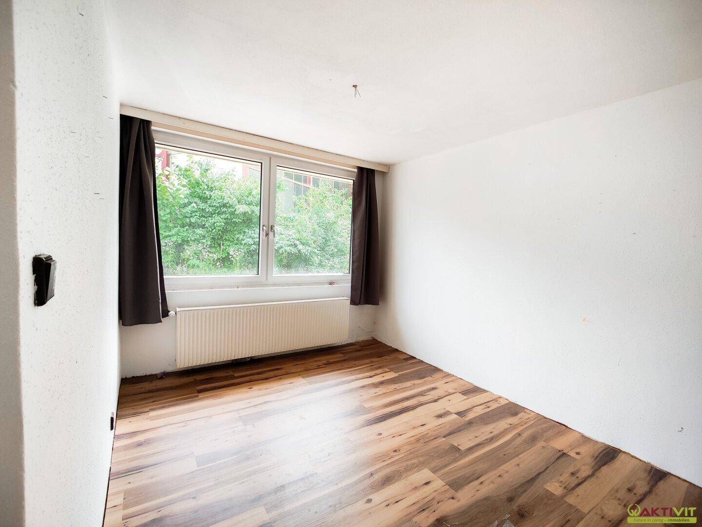 Helles Zimmer mit Laminatboden und großem Fenster, das viel Tageslicht hereinlässt.