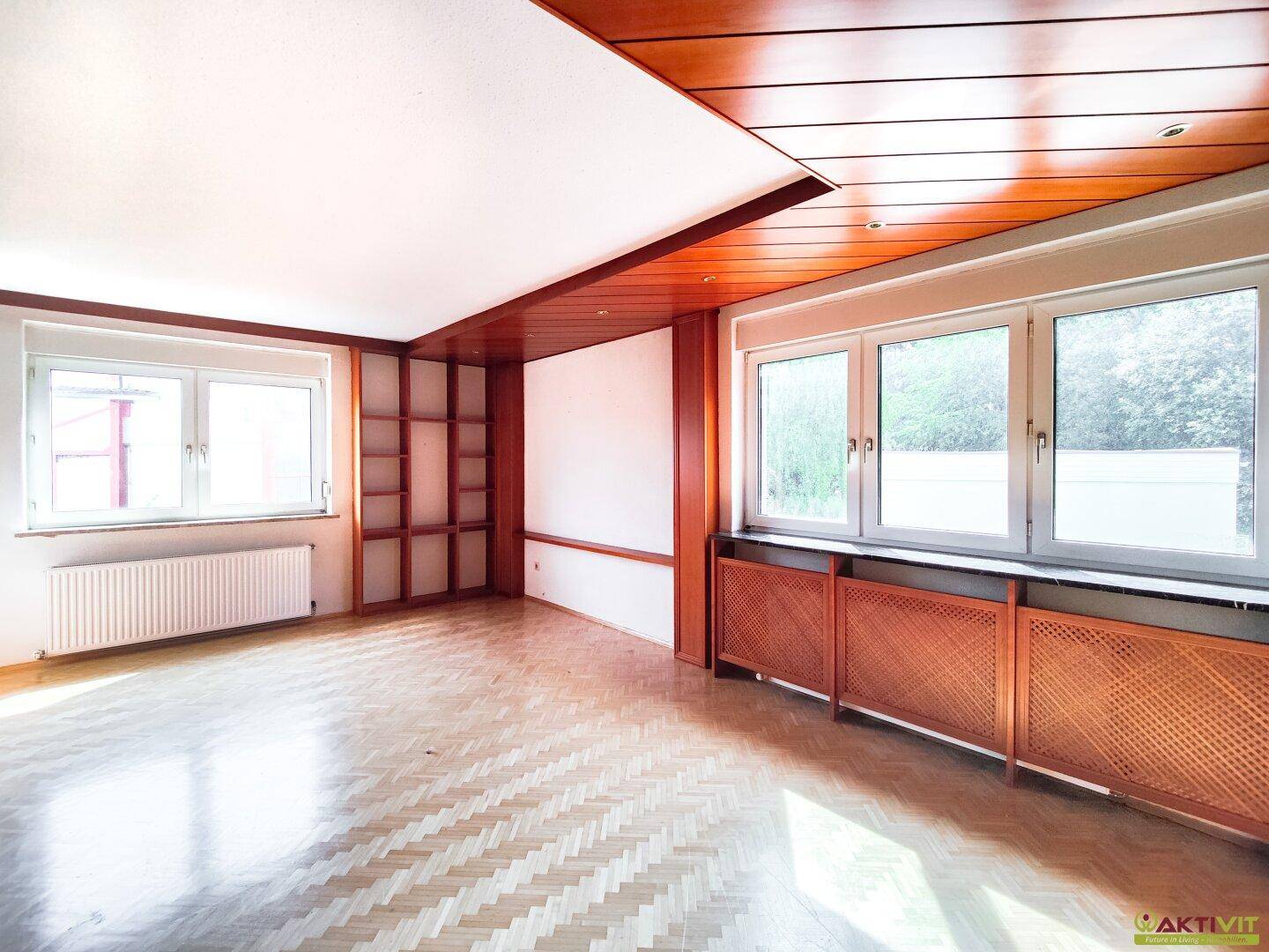 Helles Wohnzimmer mit Parkettboden, Holzvertäfelung und großen Fenstern mit Blick ins Grüne.