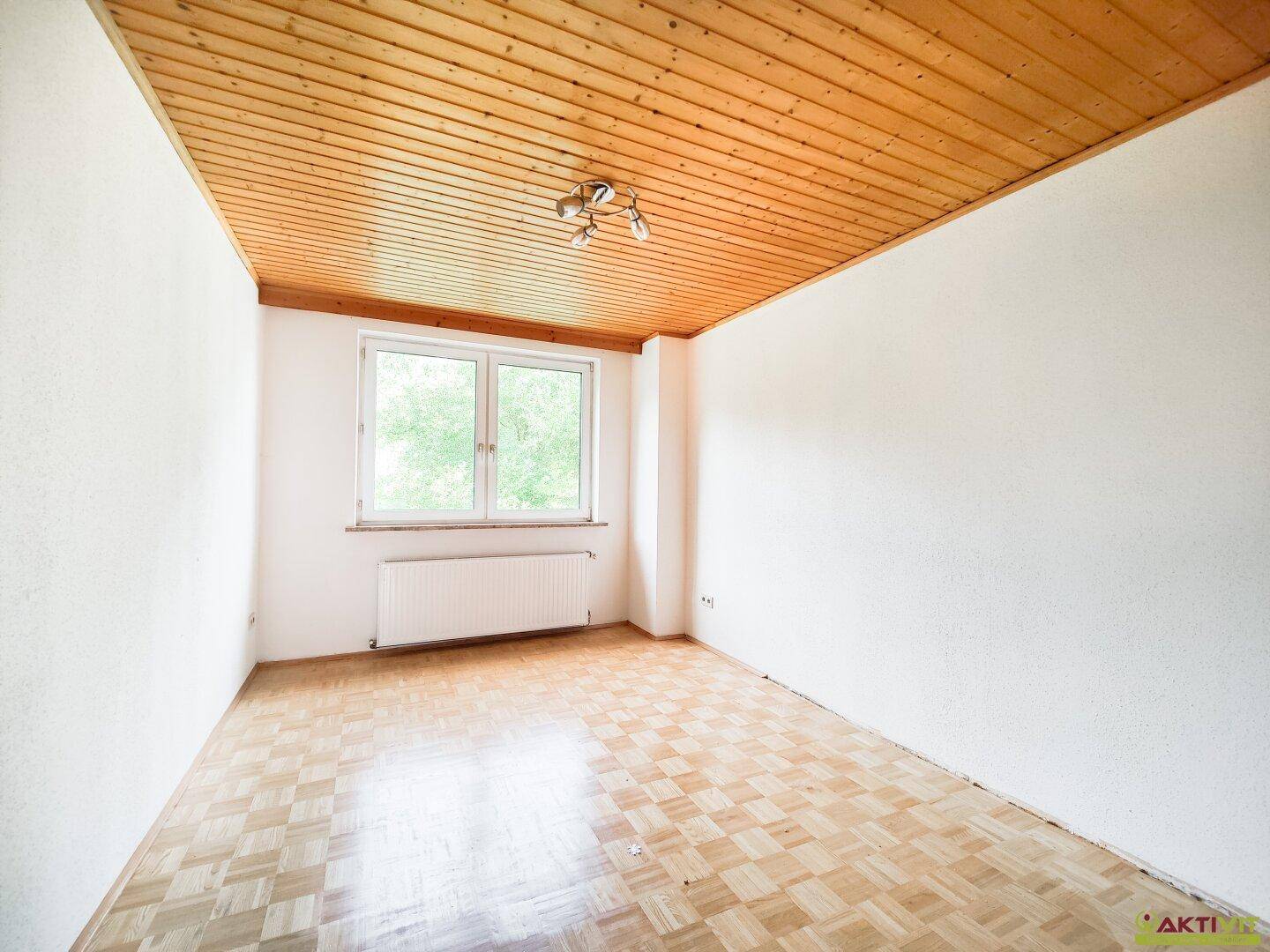 Heller Raum mit Holzdecke, Parkettboden und einem Fenster, das Tageslicht hereinlässt.