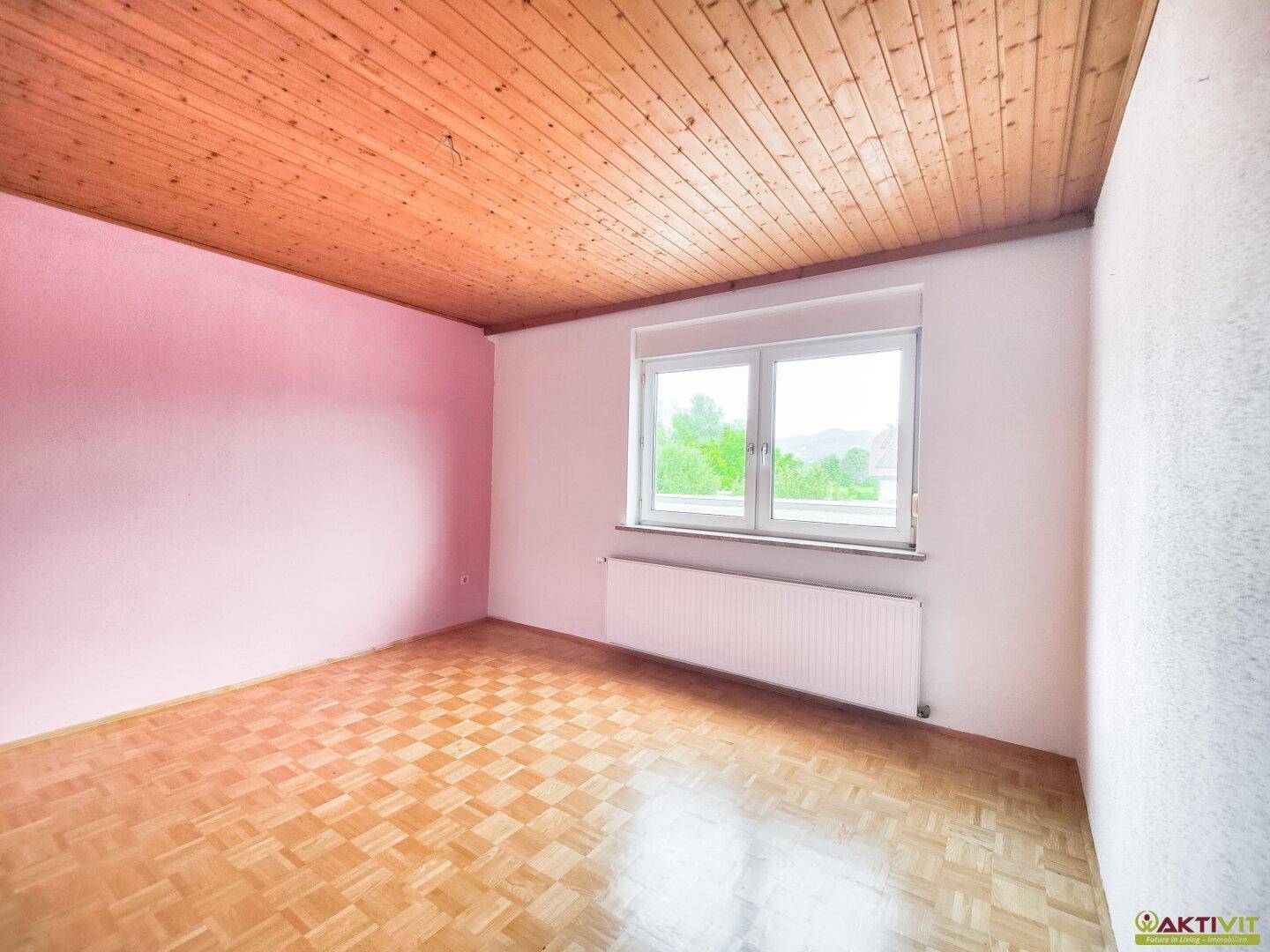 Freundlicher Raum mit Parkettboden, einer rosa Wand, Holzdecke und einem großen Fenster.