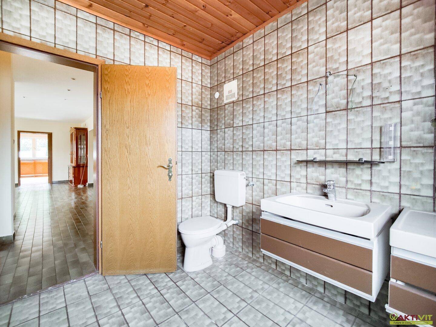 Badezimmer mit gefliesten Wänden und Boden, WC, Waschbecken und Holzdecke.