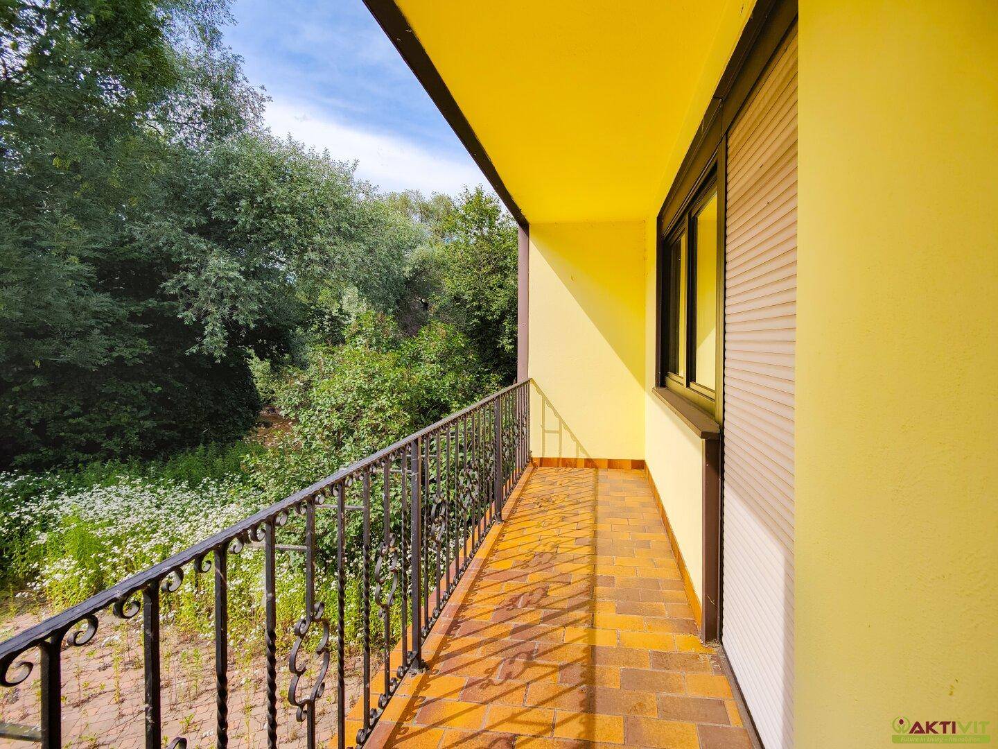 Langer Balkon mit Fliesenboden und schmiedeeisernem Geländer, umgeben von üppigem Grün.