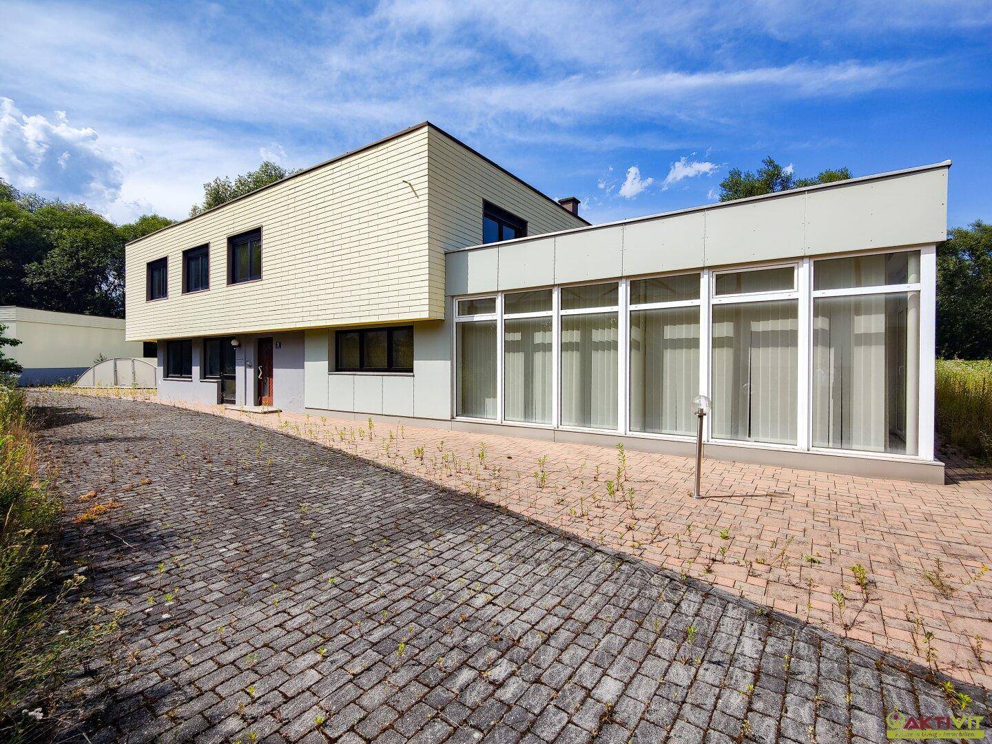 Moderne Gebäudefassade mit großen Fenstern und gepflastertem Vorplatz, umgeben von Natur.