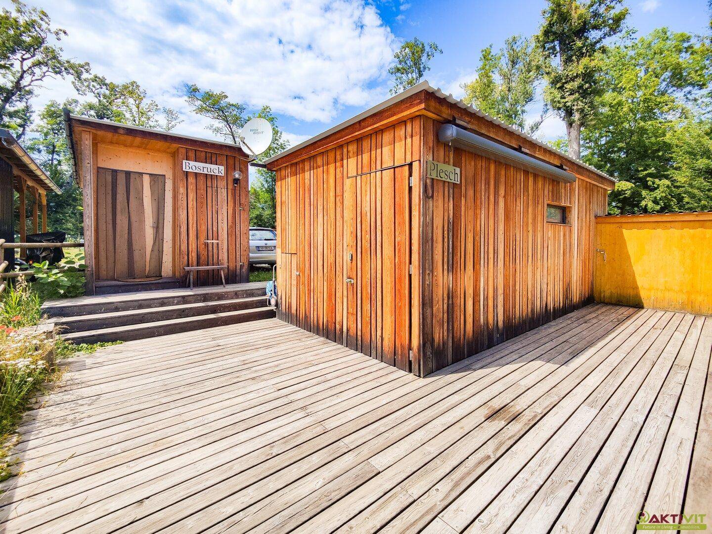 Zwei kleine Holzhütten mit einer Holzterrasse davor unter strahlend blauem Himmel.