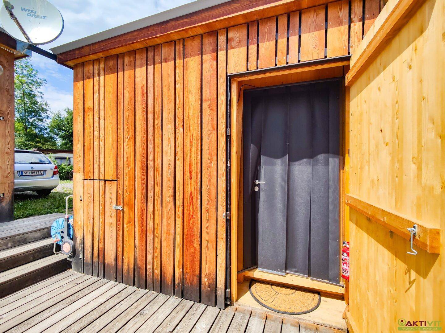 Offene Holztür einer kleinen Hütte mit Holzfassade und Satellitenschüssel an der Seite.