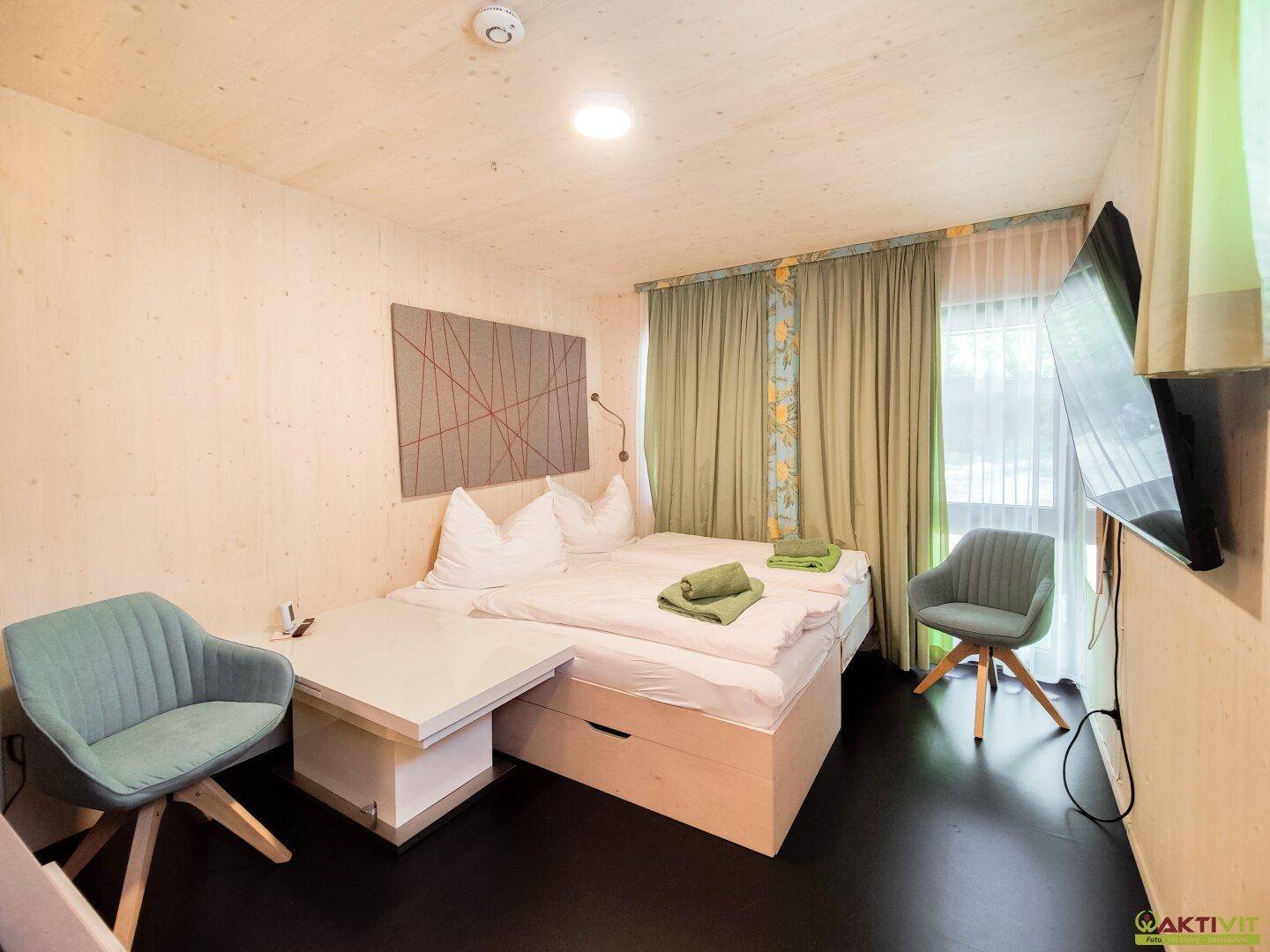 Gemütliches Schlafzimmer mit Doppelbett, zwei Stühlen und einem an der Wand montierten Fernseher.