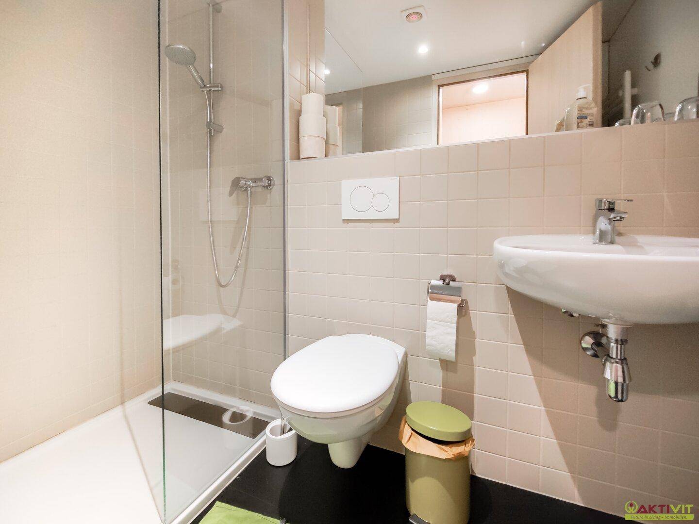 Modernes Badezimmer mit Duschkabine, Toilette und kleinem Waschbecken in hellen Fliesen.