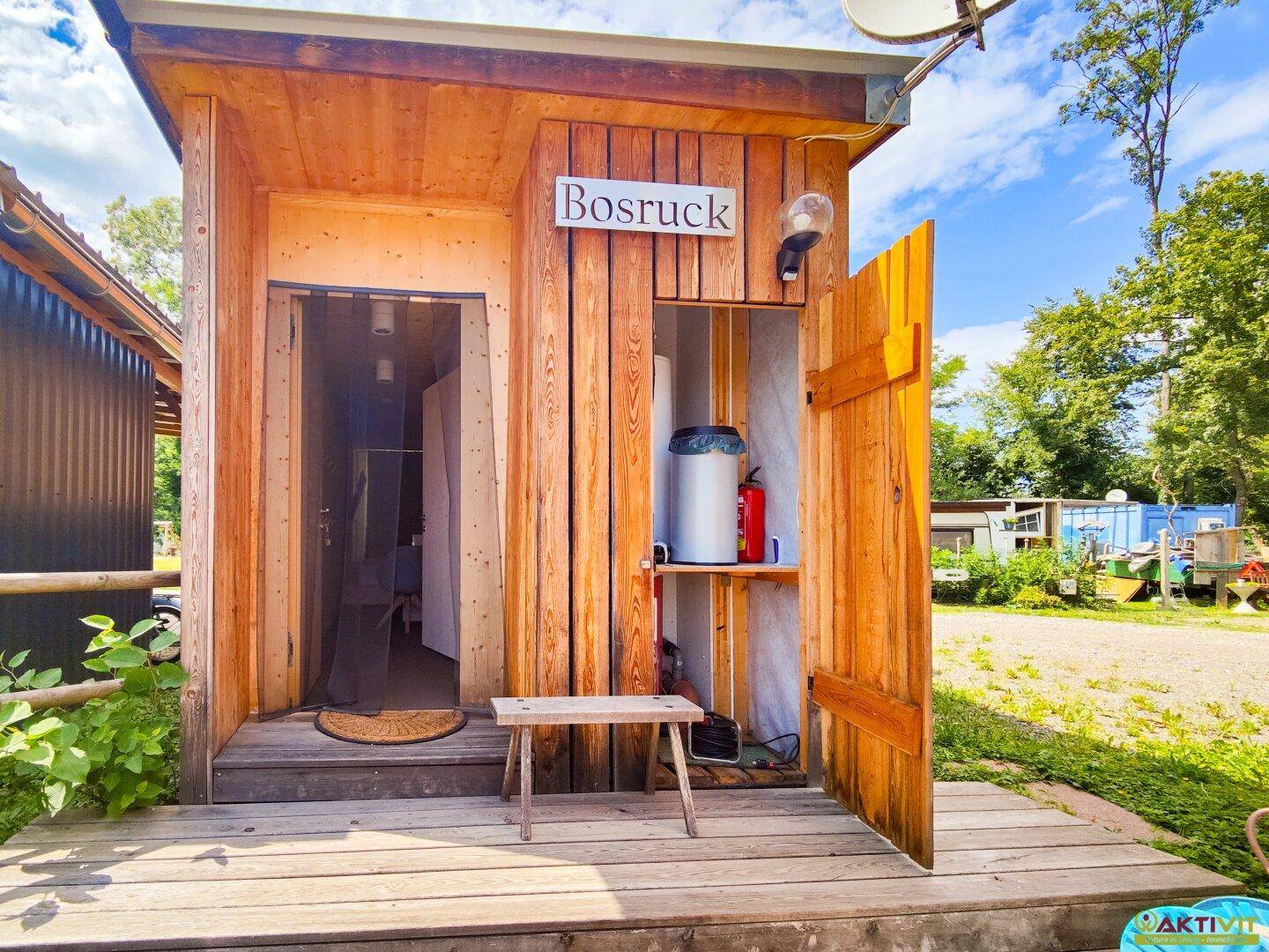 Eingangsbereich einer Holzhütte mit offener Tür und einem kleinen Holzbänkchen davor.