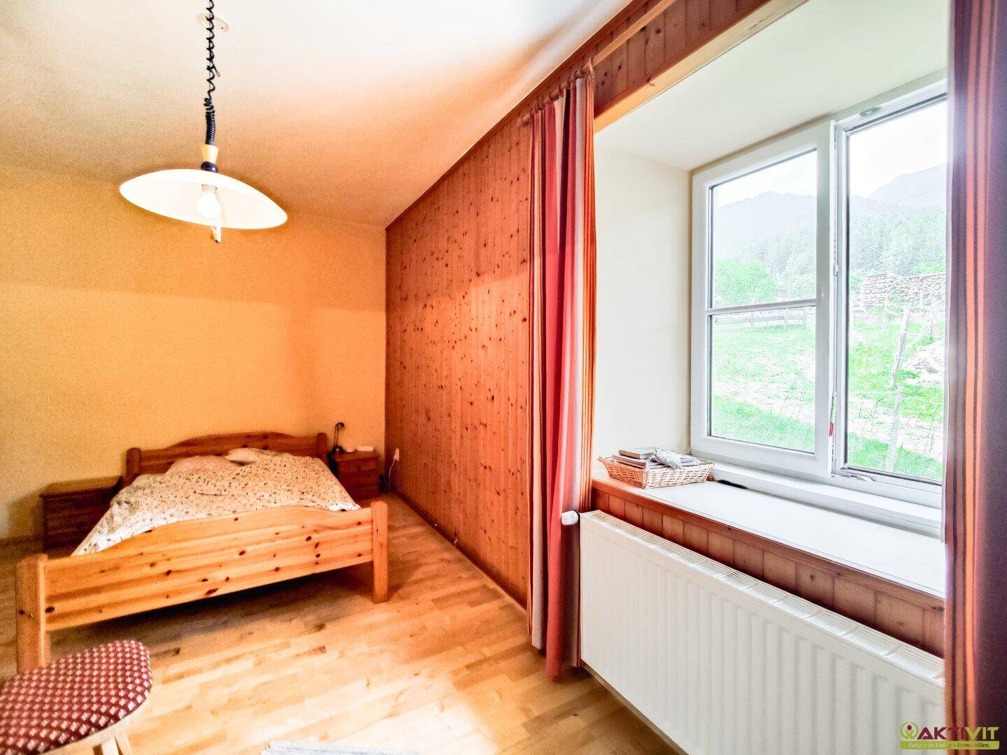 Schlafzimmer mit Holzbett, Holzvertäfelung und großem Fenster, das viel Tageslicht hereinlässt.