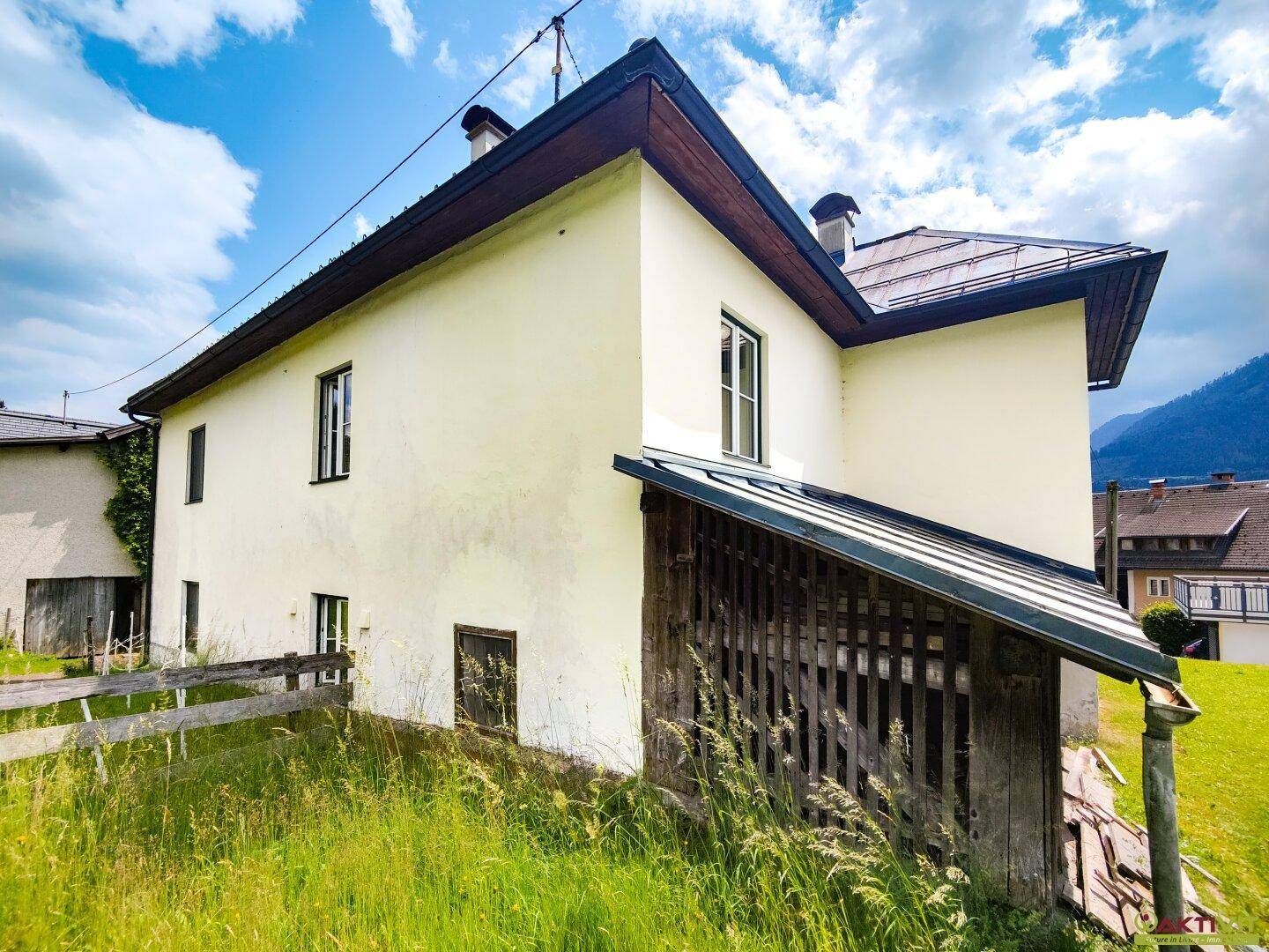 Seitenansicht des Hauses mit einem rustikalen Holzunterstand und grüner Wiese.
