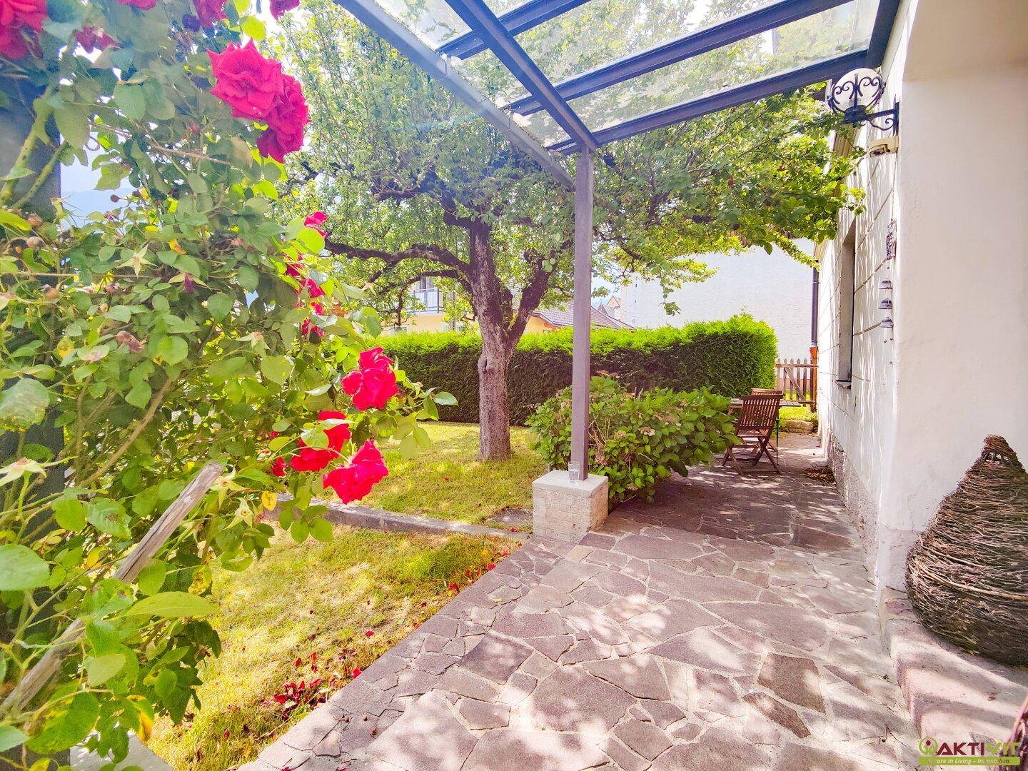 Überdachte Terrasse mit Steinboden, blühenden Rosen und Blick in den grünen Garten.