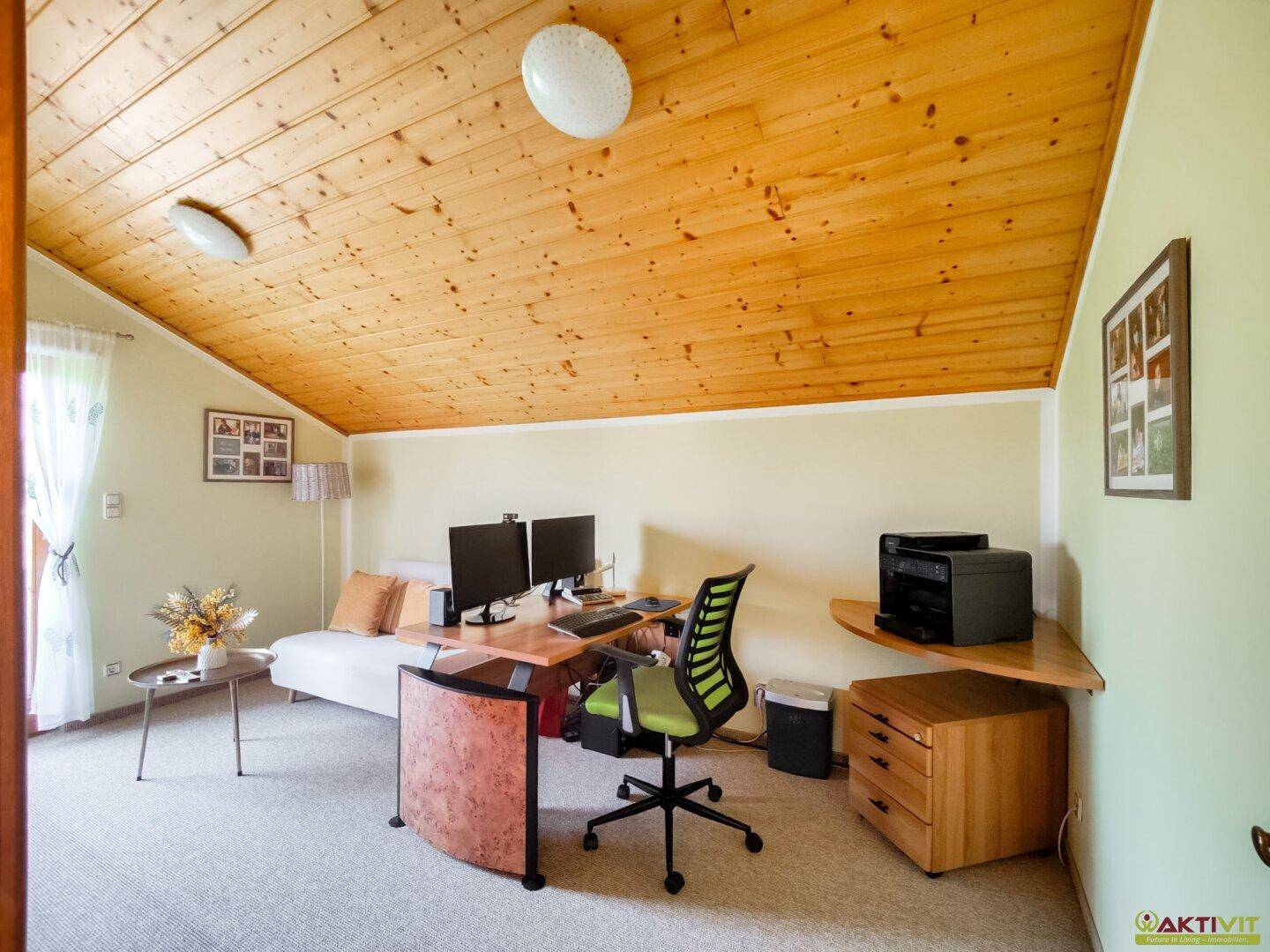 Helles Arbeitszimmer mit Dachschräge, Teppichboden und einem großen Schreibtisch mit Computern.