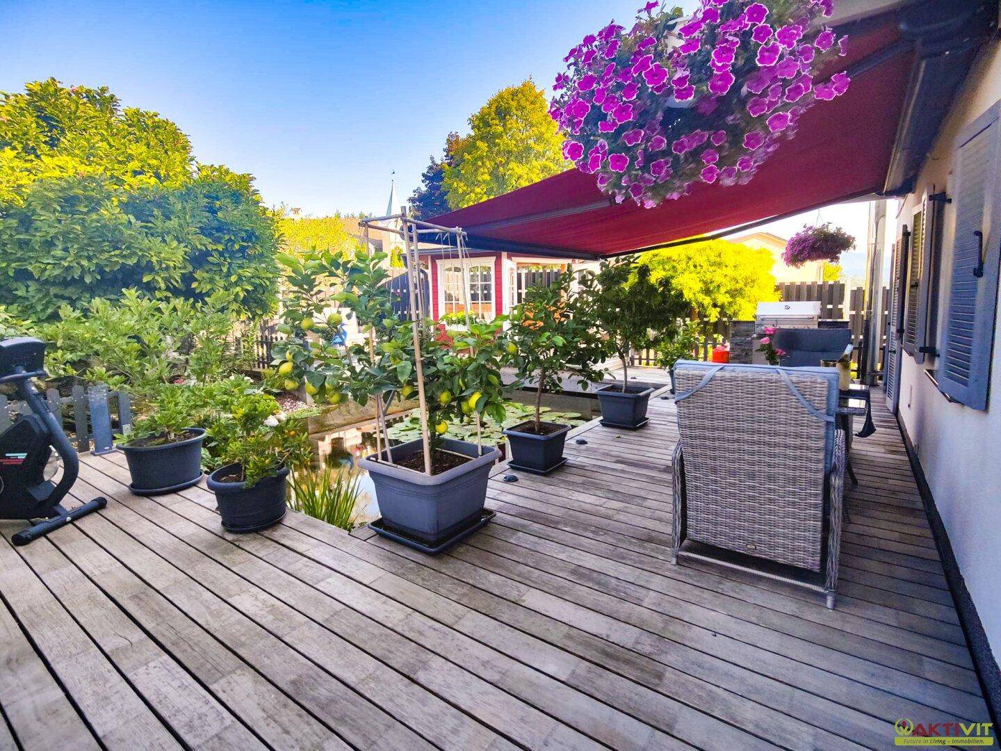 Gemütliche Holzterrasse mit Gartenmöbeln, Zierpflanzen, einem Teich und einer roten Markise.