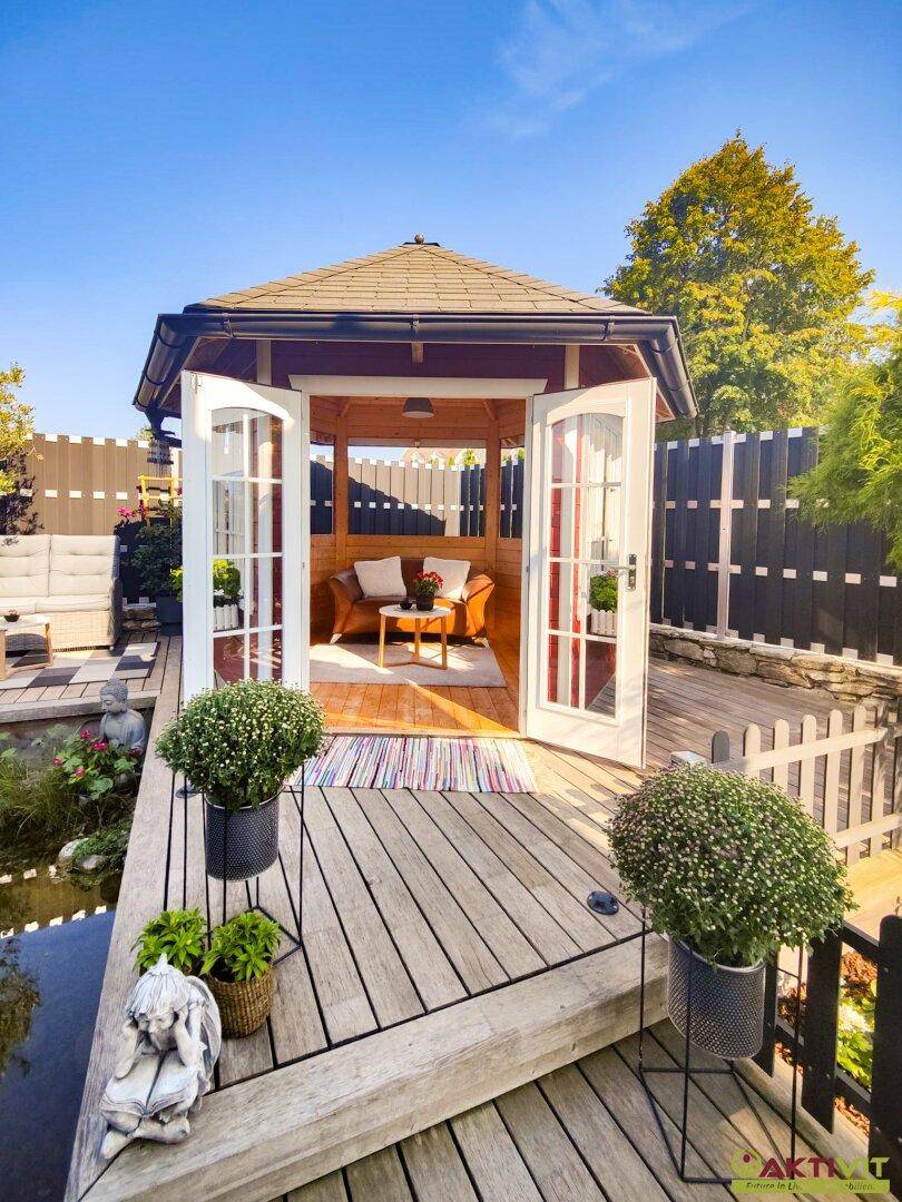 Charmantes Holz-Gartenhaus mit geöffneten Türen, umgeben von einer Holzterrasse und einem Teich.