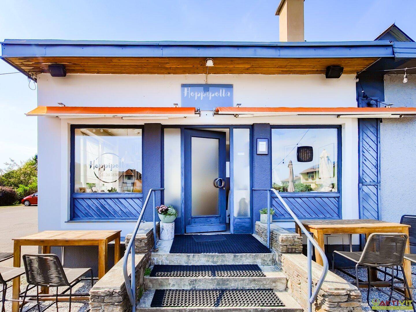 Eingangsbereich eines Gebäudes mit blauer Fassade und Holzdetails, führt zu einem Café oder Restaurant.