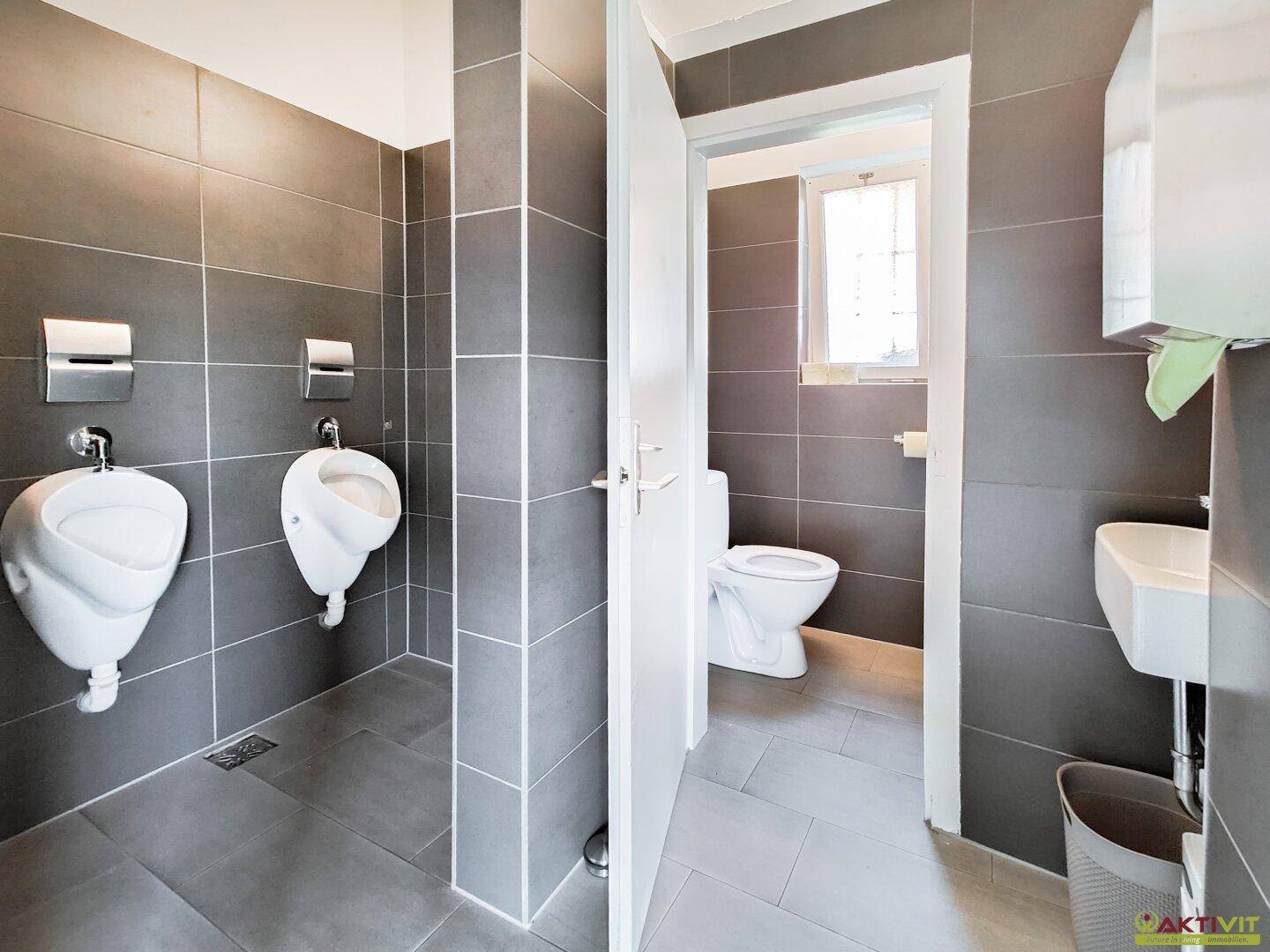 Moderne öffentliche Toilettenanlage mit grauen Fliesen, Urinalen und separater WC-Kabine.