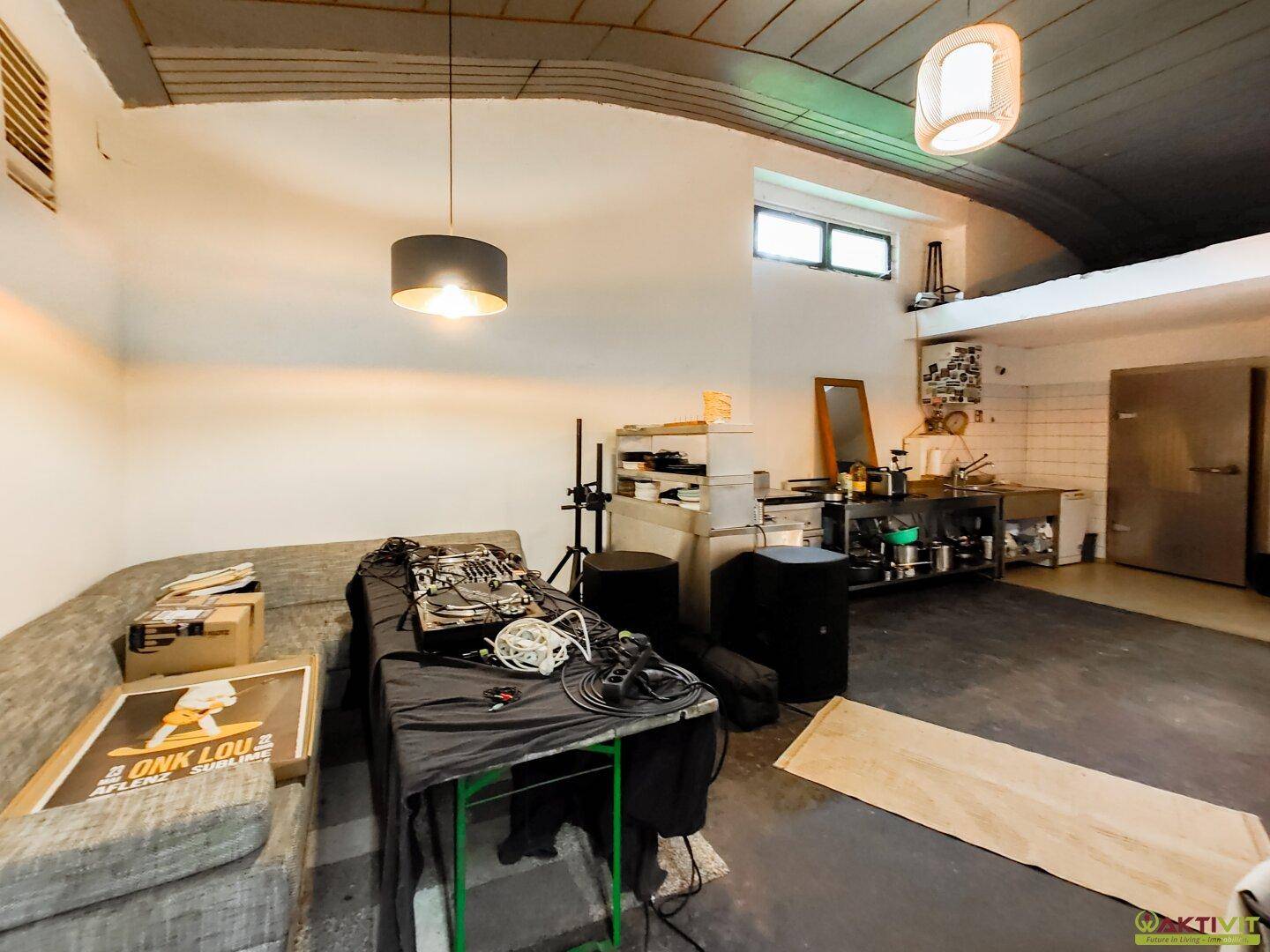 Geräumiger Raum mit Sofa, Veranstaltungstechnik und einer kleinen Küchenzeile im Hintergrund, unter einer rohen Deckenkonstruktion.
