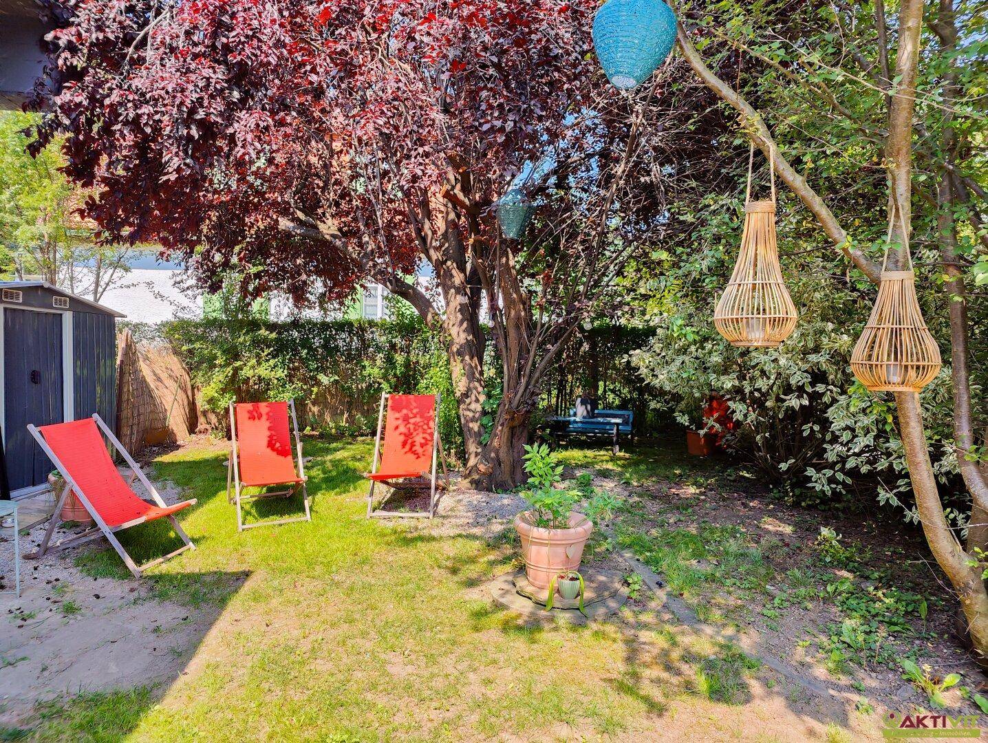 Idyllischer Gartenbereich mit einem Baum mit roten Blättern, roten Liegestühlen und dekorativen Laternen.