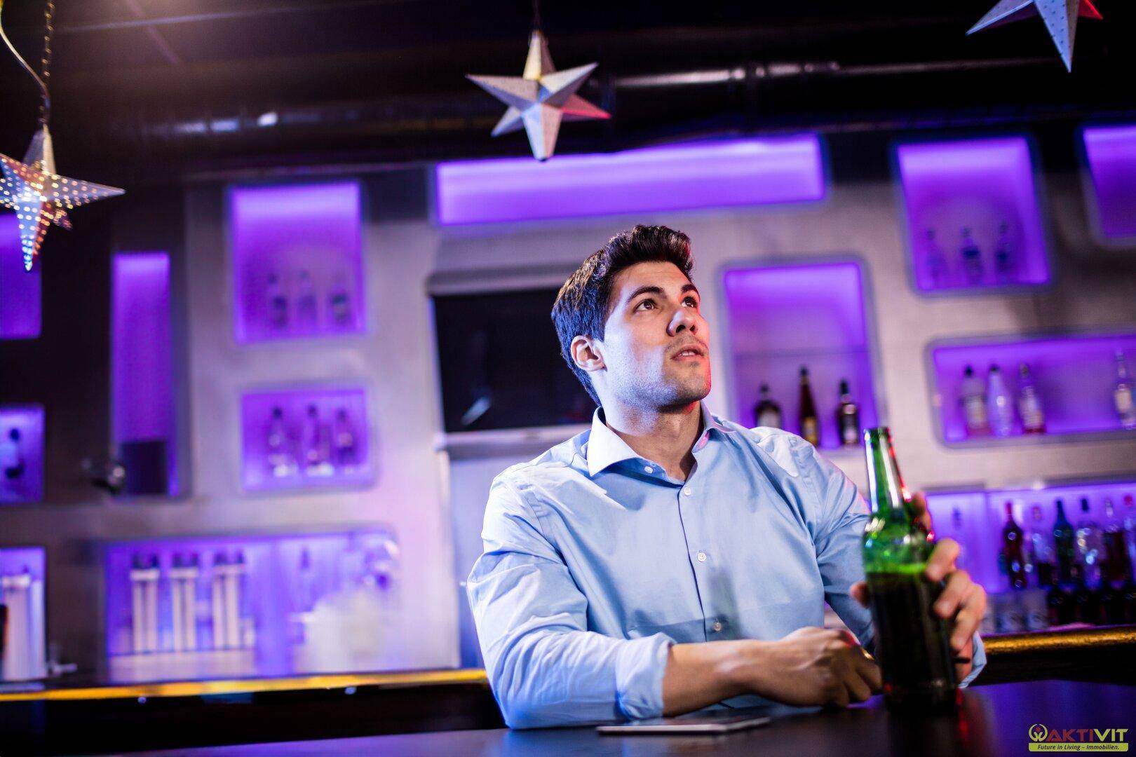 Mann sitzt an einer Bar mit lila beleuchteten Regalen und Sternendekoration an der Decke.