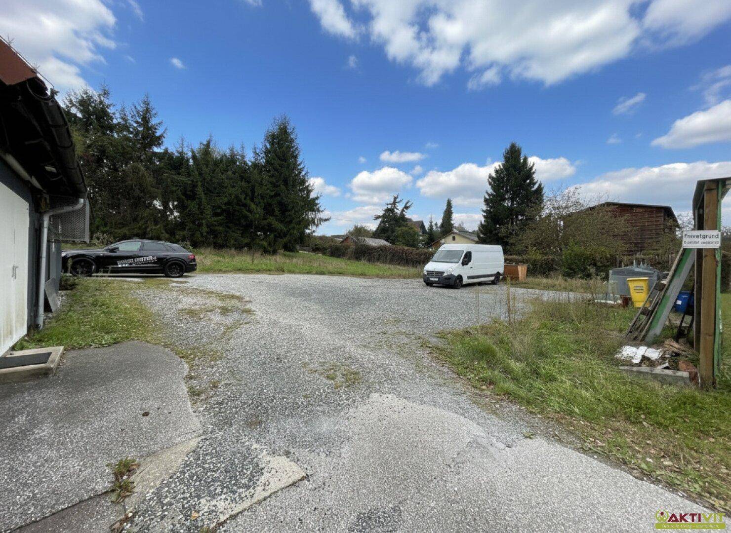 Großer Schotterparkplatz mit mehreren Fahrzeugen und Bäumen am Rand des Grundstücks unter blauem Himmel.