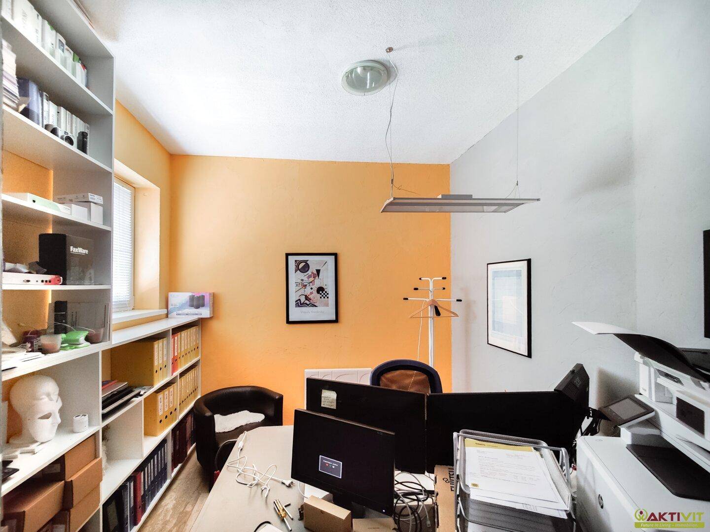 Helles Büro mit orangefarbener Akzentwand, Regalen und zwei Arbeitsplätzen.