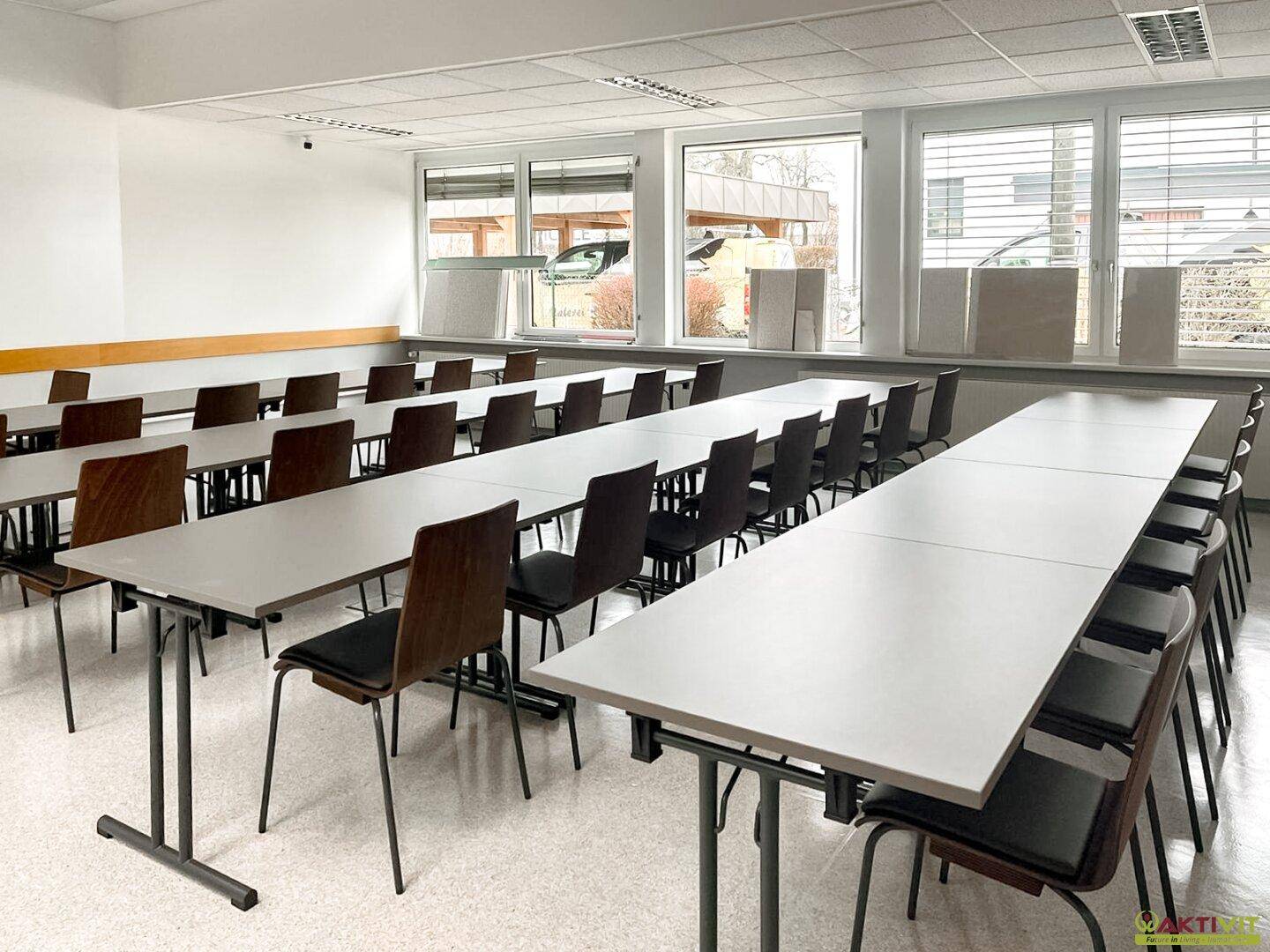 Großer Schulungsraum mit vielen Tischen und Stühlen, hell beleuchtet durch Fenster, ideal für Unterricht oder Konferenzen.