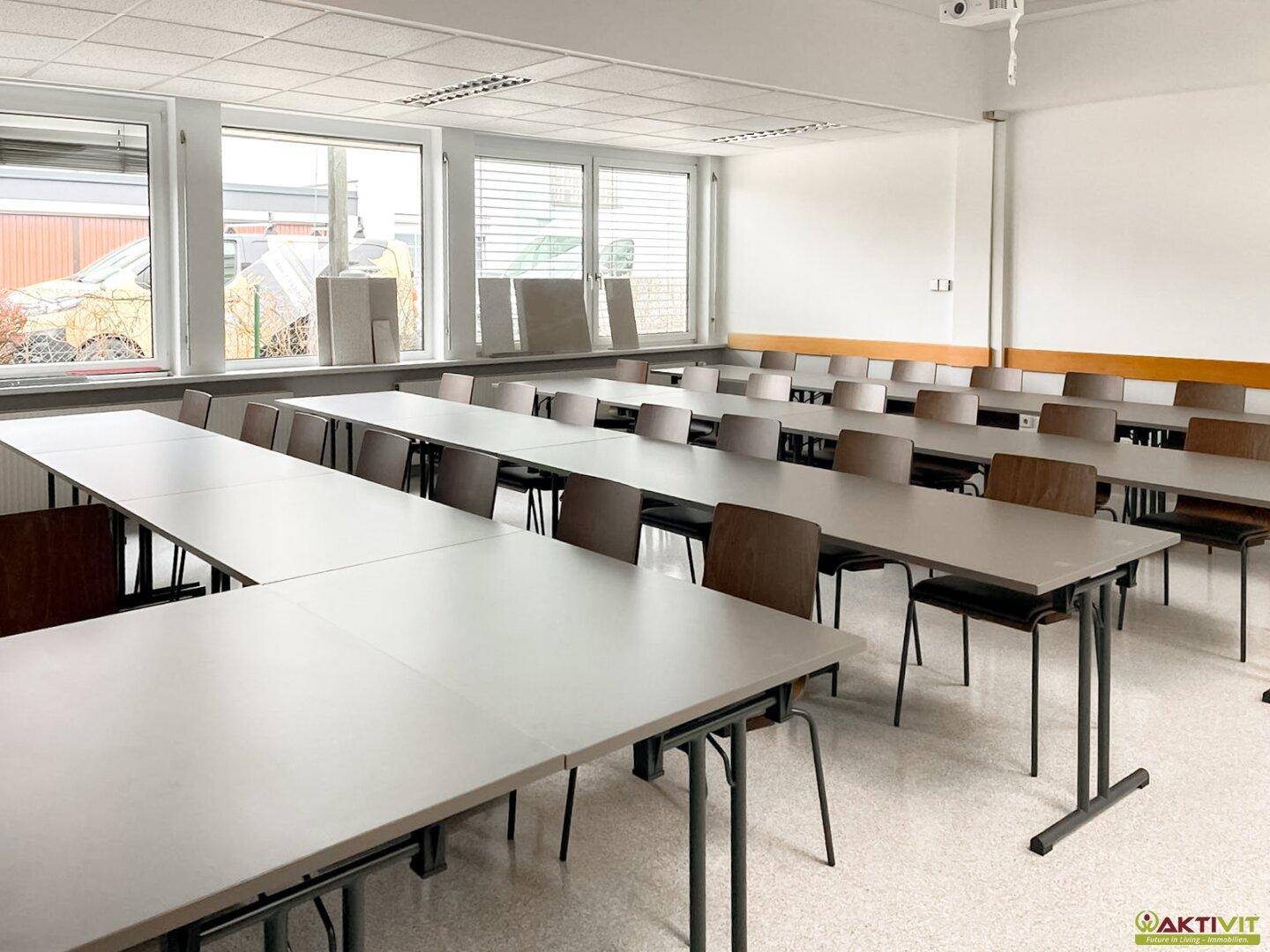 Geräumiger Schulungsraum mit Reihen von Tischen und Stühlen, hell und funktional, mit großen Fenstern und Fliesenboden.