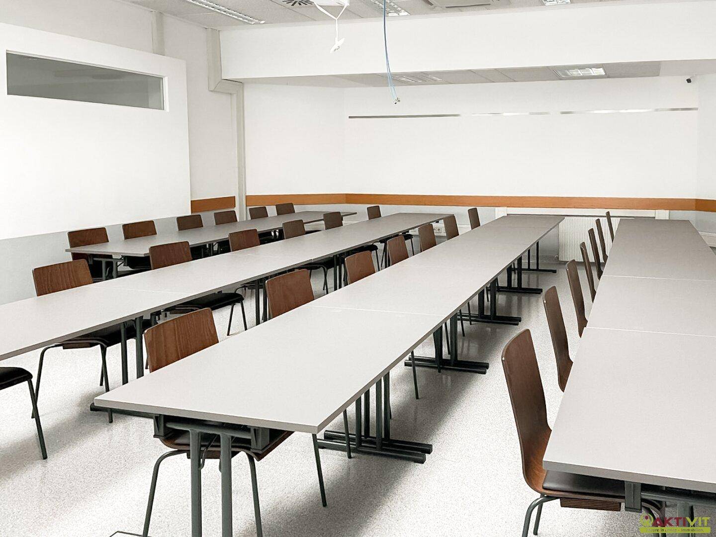 Ordentlicher Schulungsraum mit Reihen von Tischen und Stühlen, hellem Holzfußboden und schlichtem, funktionalem Design.