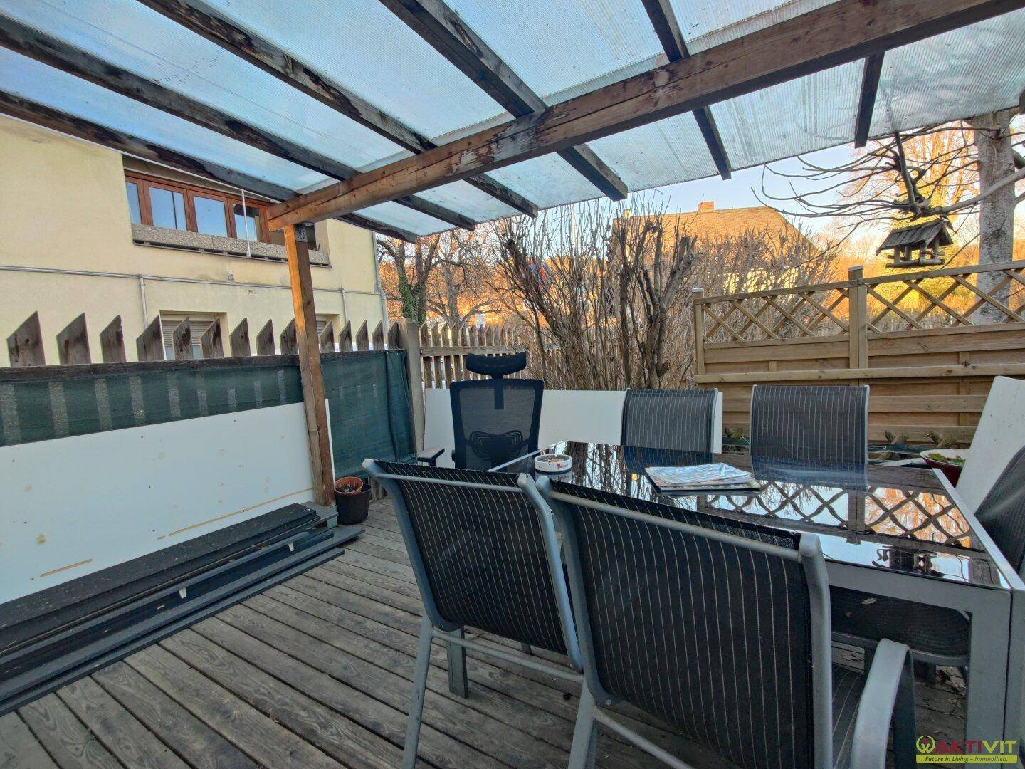 Überdachte Holzterrasse mit Gartenmöbeln und Blick auf den umzäunten Außenbereich mit Bäumen.