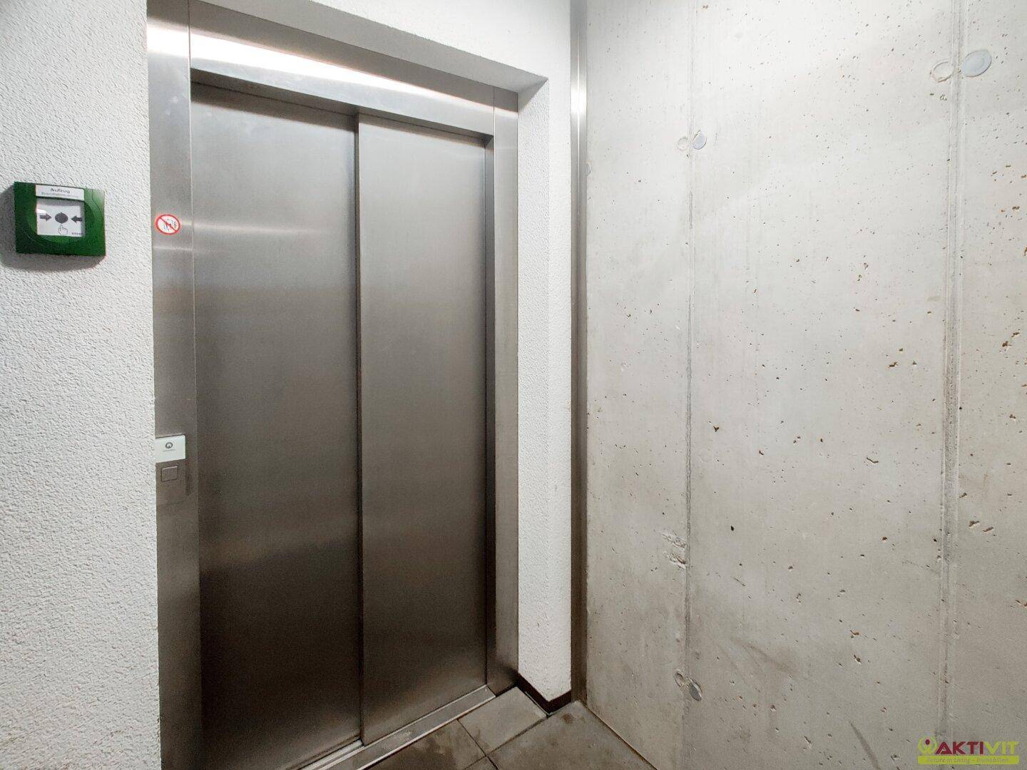 Moderner Aufzug mit Edelstahltüren in einem hellen Flur mit Betonwand.
